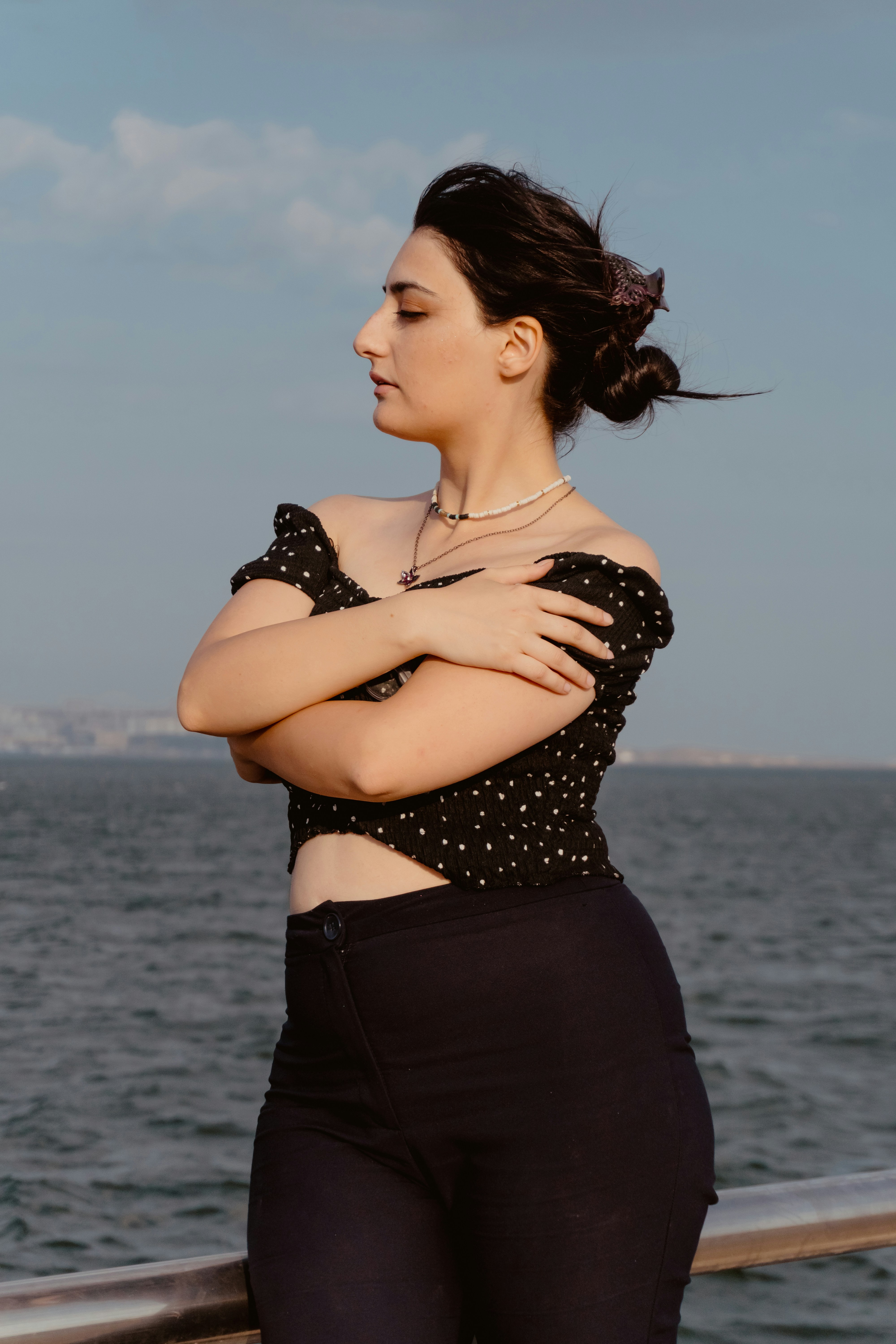 A woman with hair in a bun standing by the sea with arms crossed