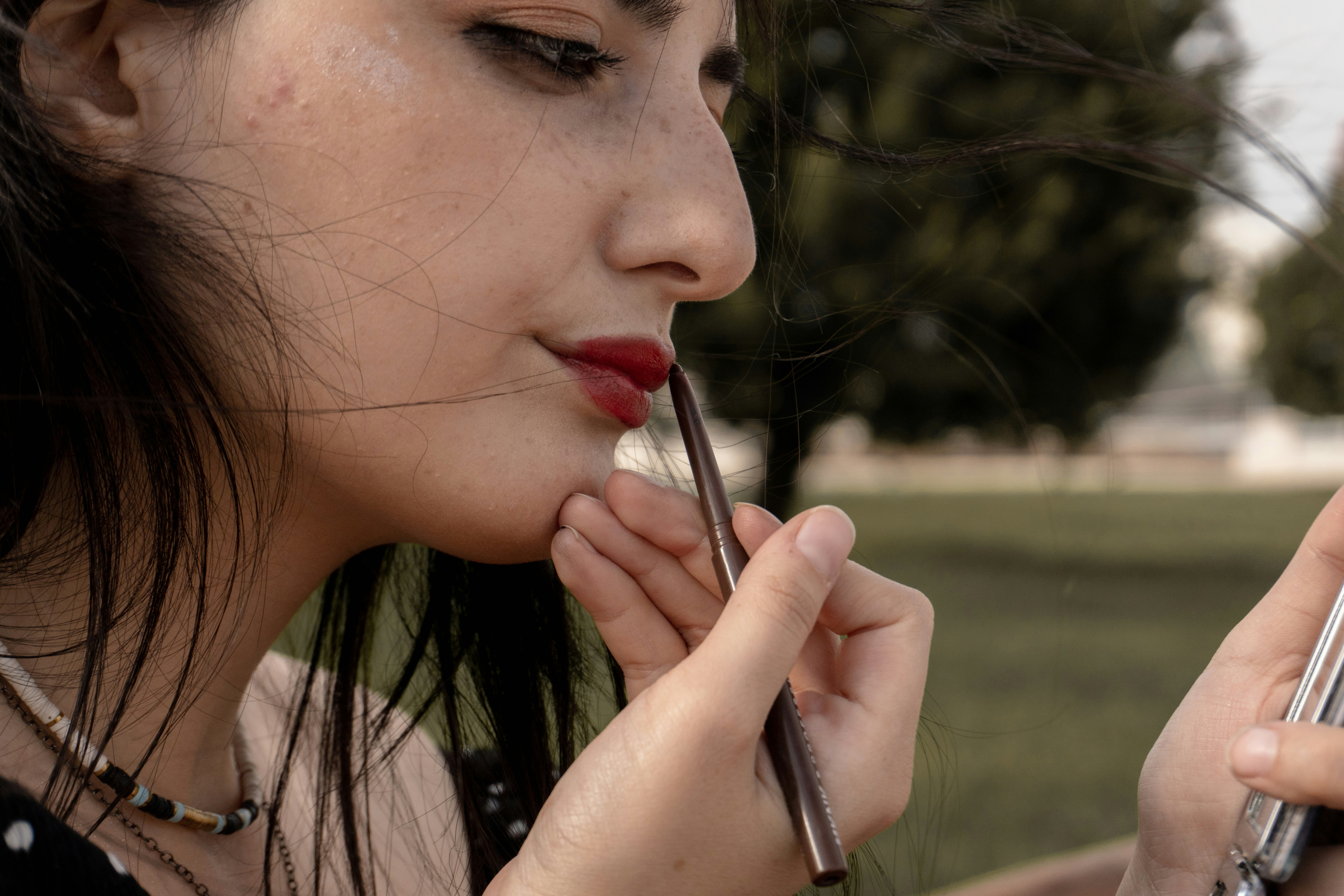 A woman applying red lip liner in a mirror outdoors