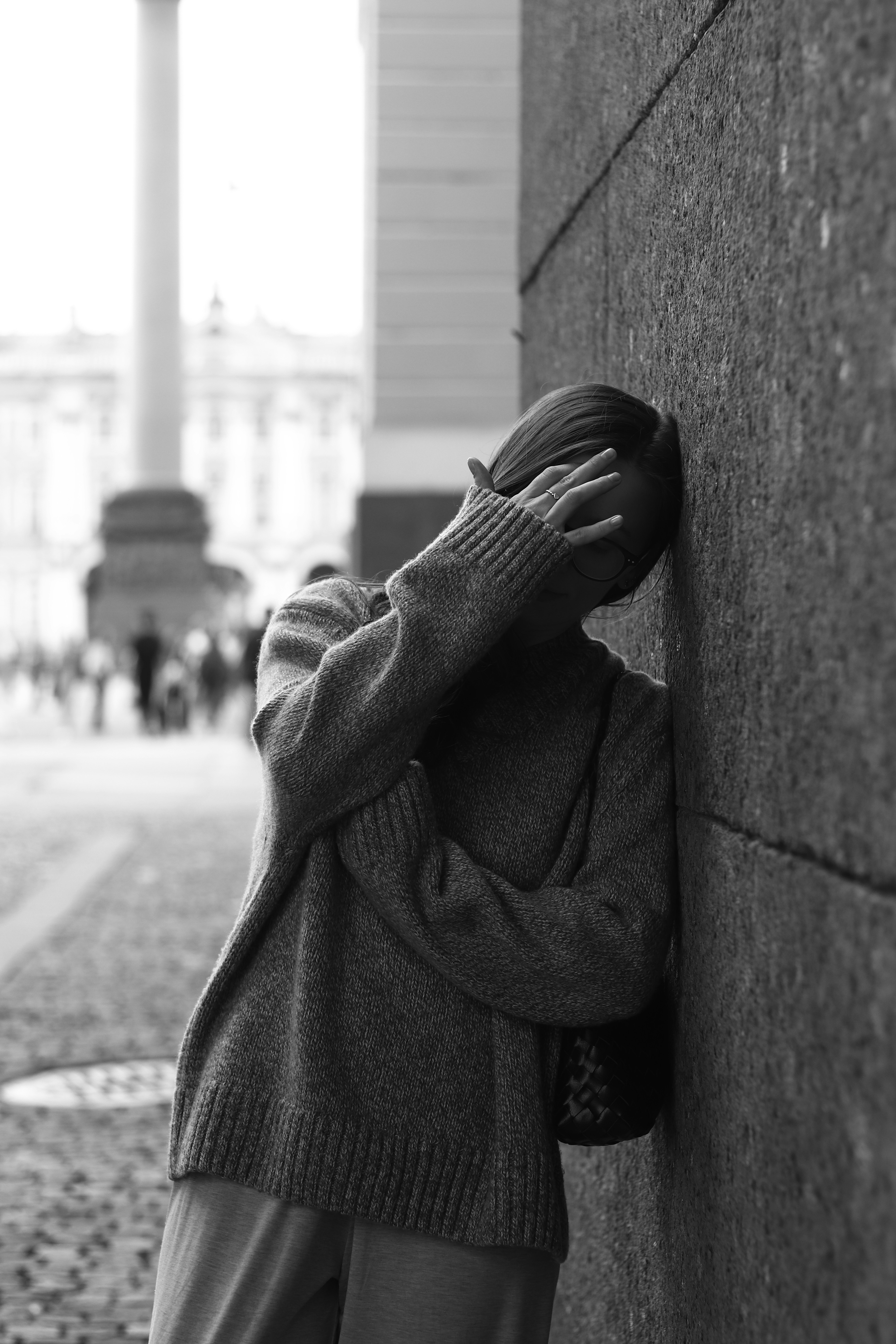 A person in a knit sweater leaning against a stone wall on a city street