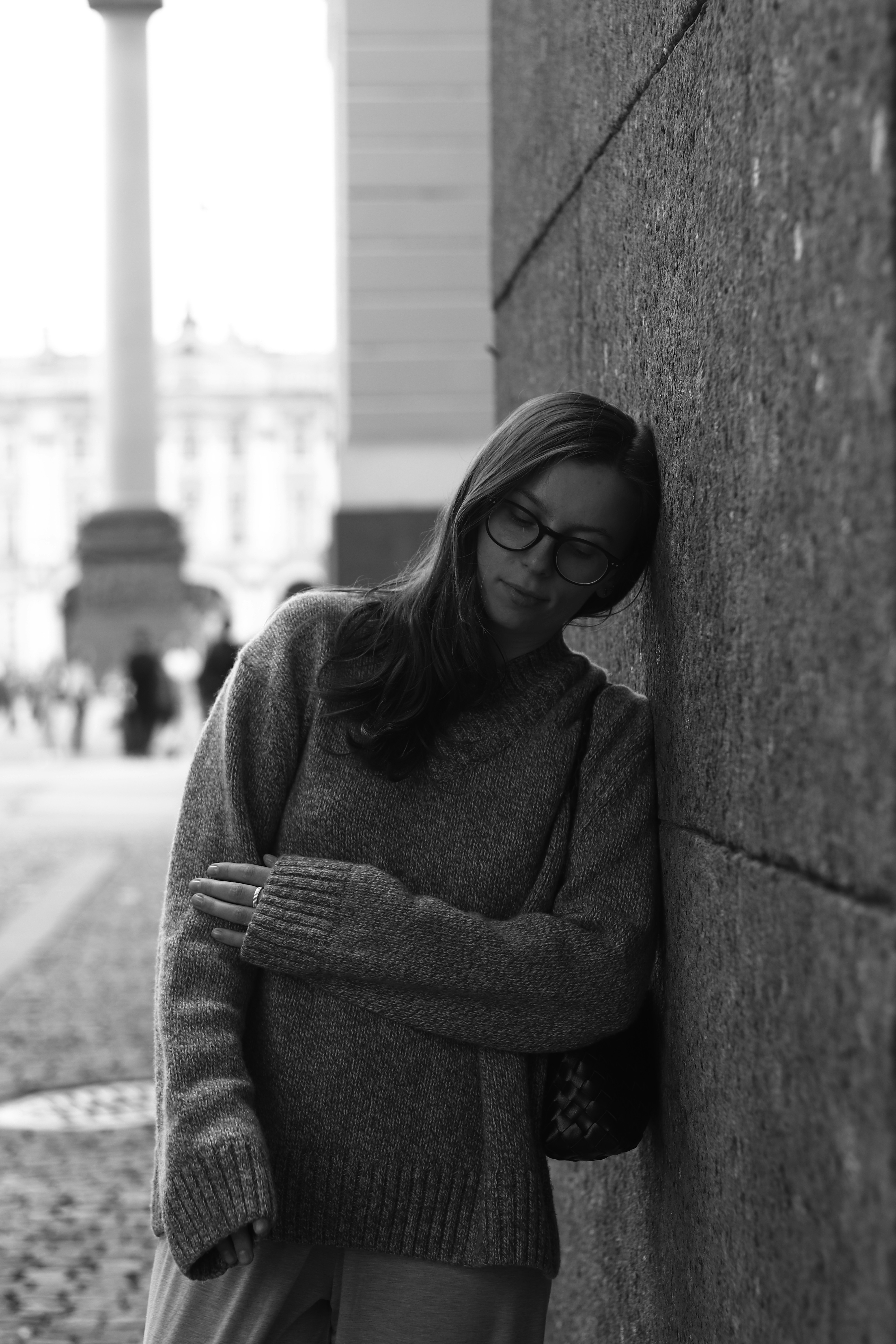 A woman in glasses and a sweater leaning against a stone wall outdoors