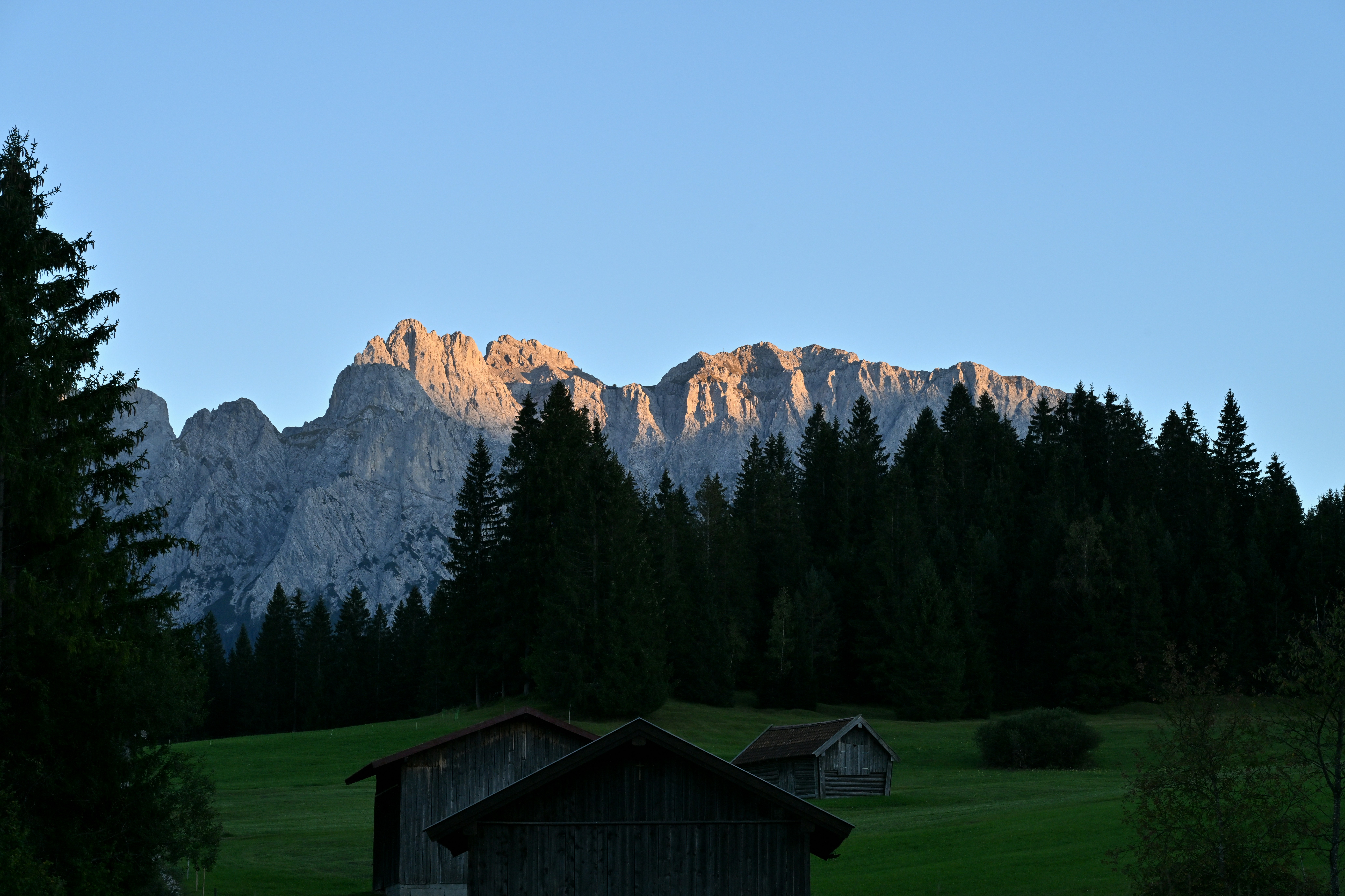 Wooden huts in a green meadow before a forest and sunlit mountain peaks