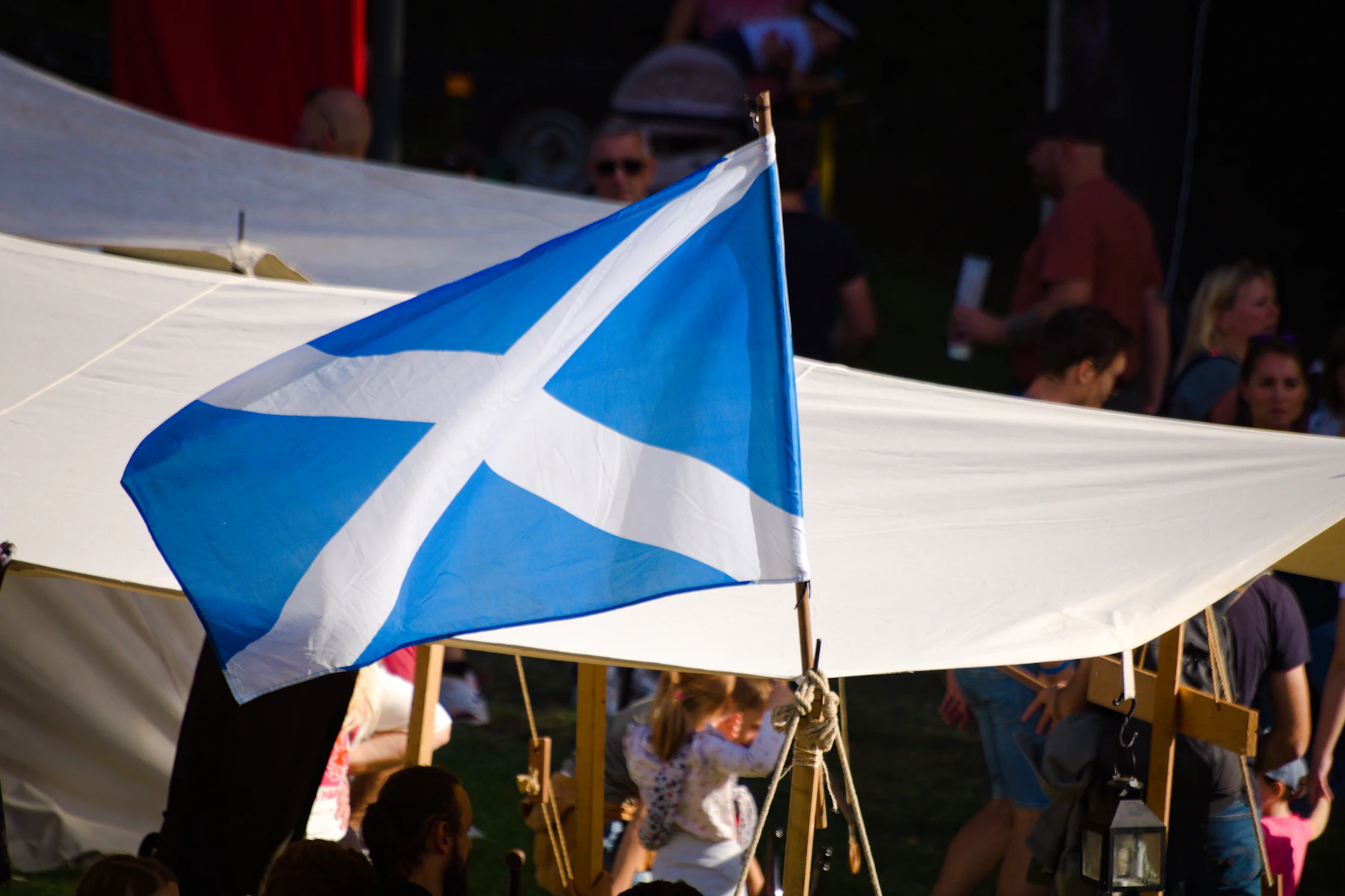 A blue and white saltire flag flying beside a white canvas tent, with a festival crowd in the background