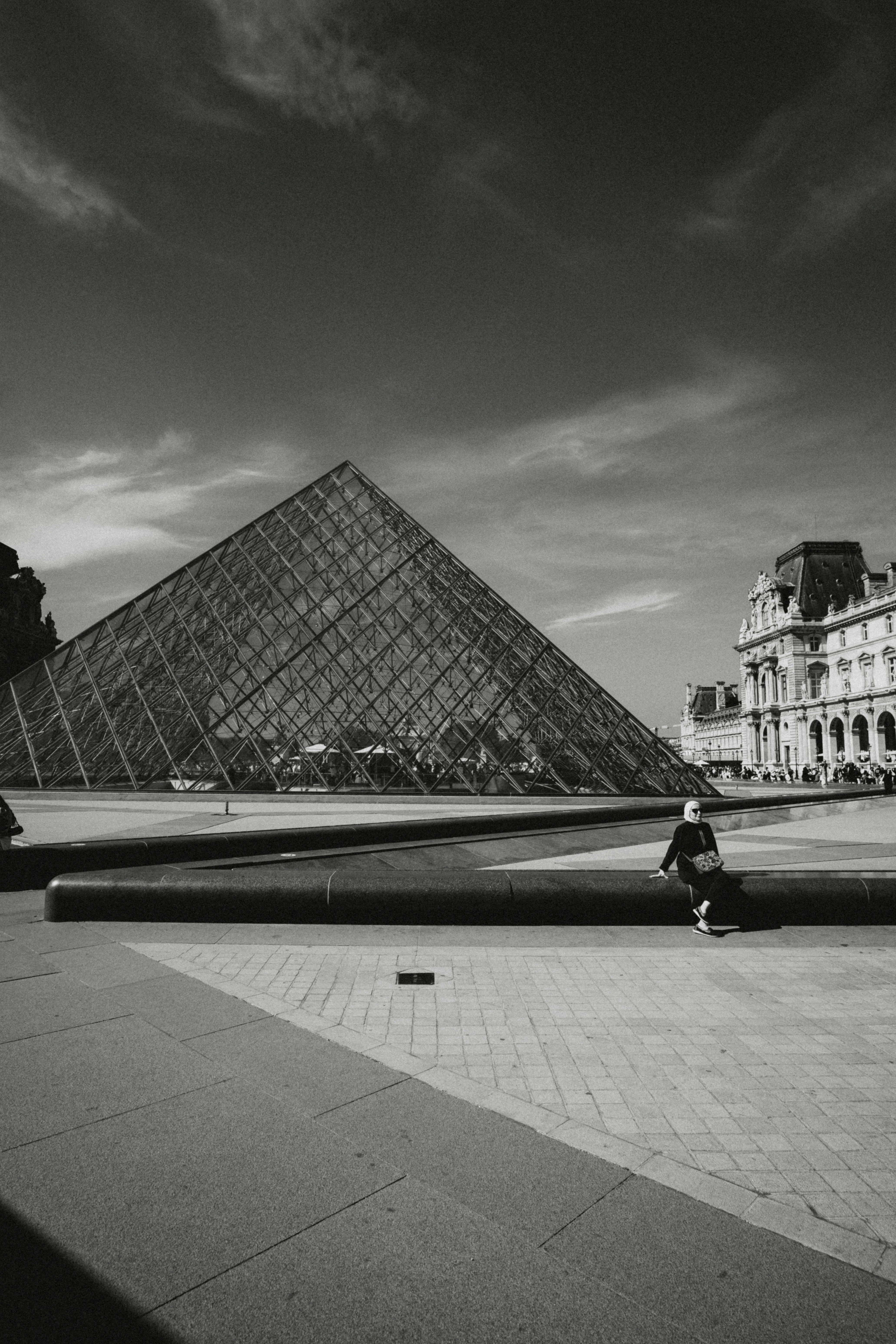 A person in a headscarf sitting by the Louvre Pyramid in Paris