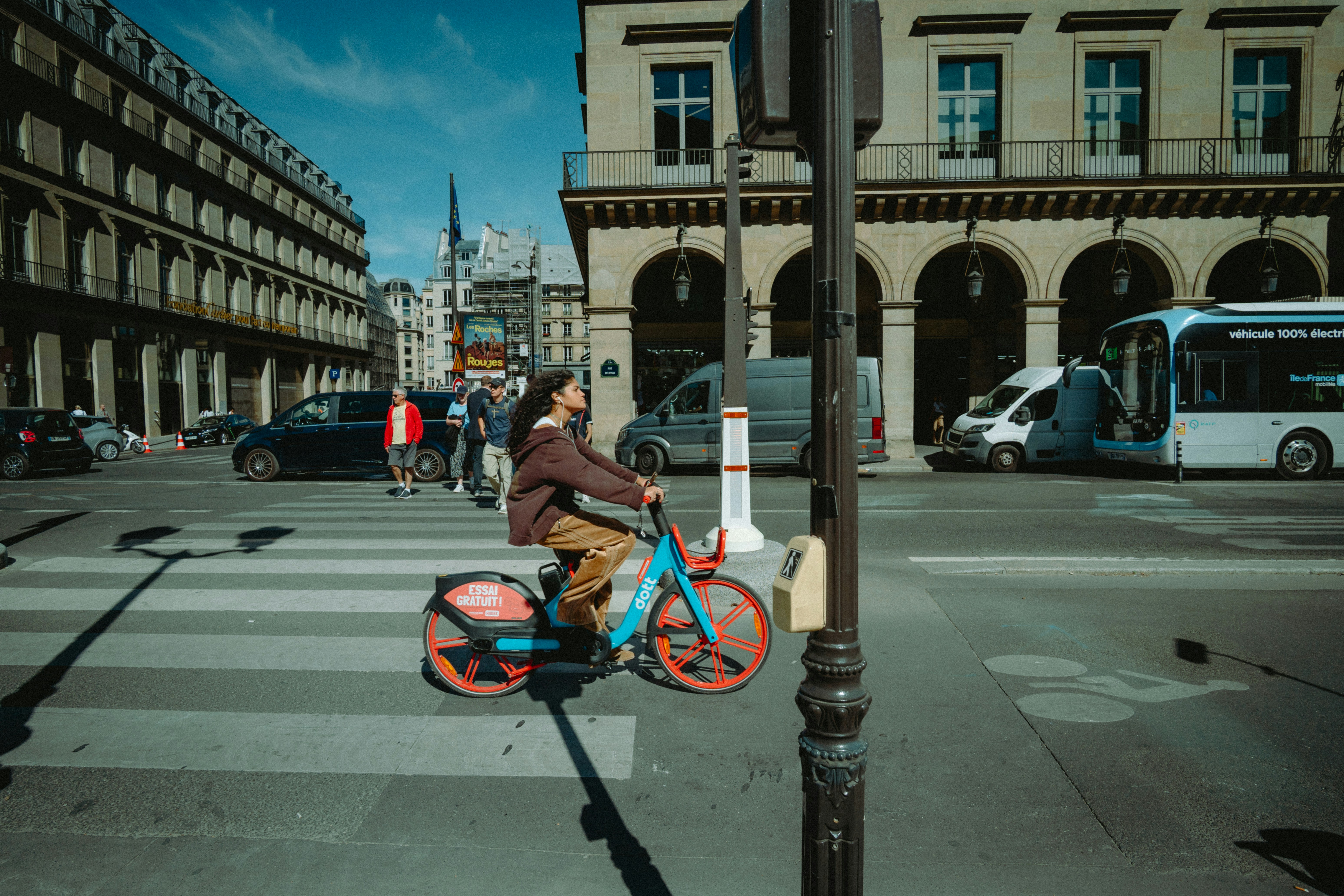 A person cycling a blue and orange rental bike across a Paris street crossing