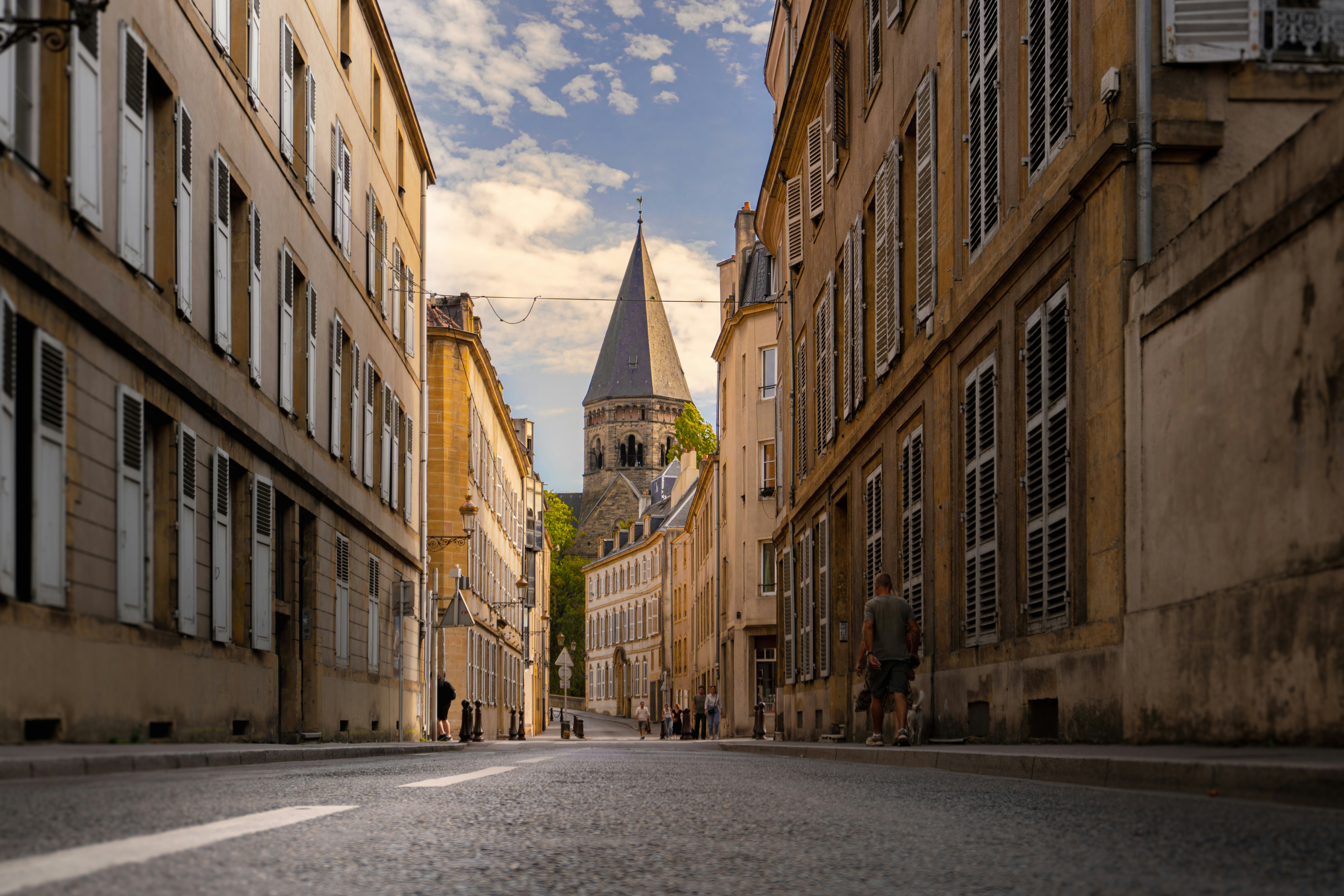 A person walking a dog down a narrow street toward a stone church tower