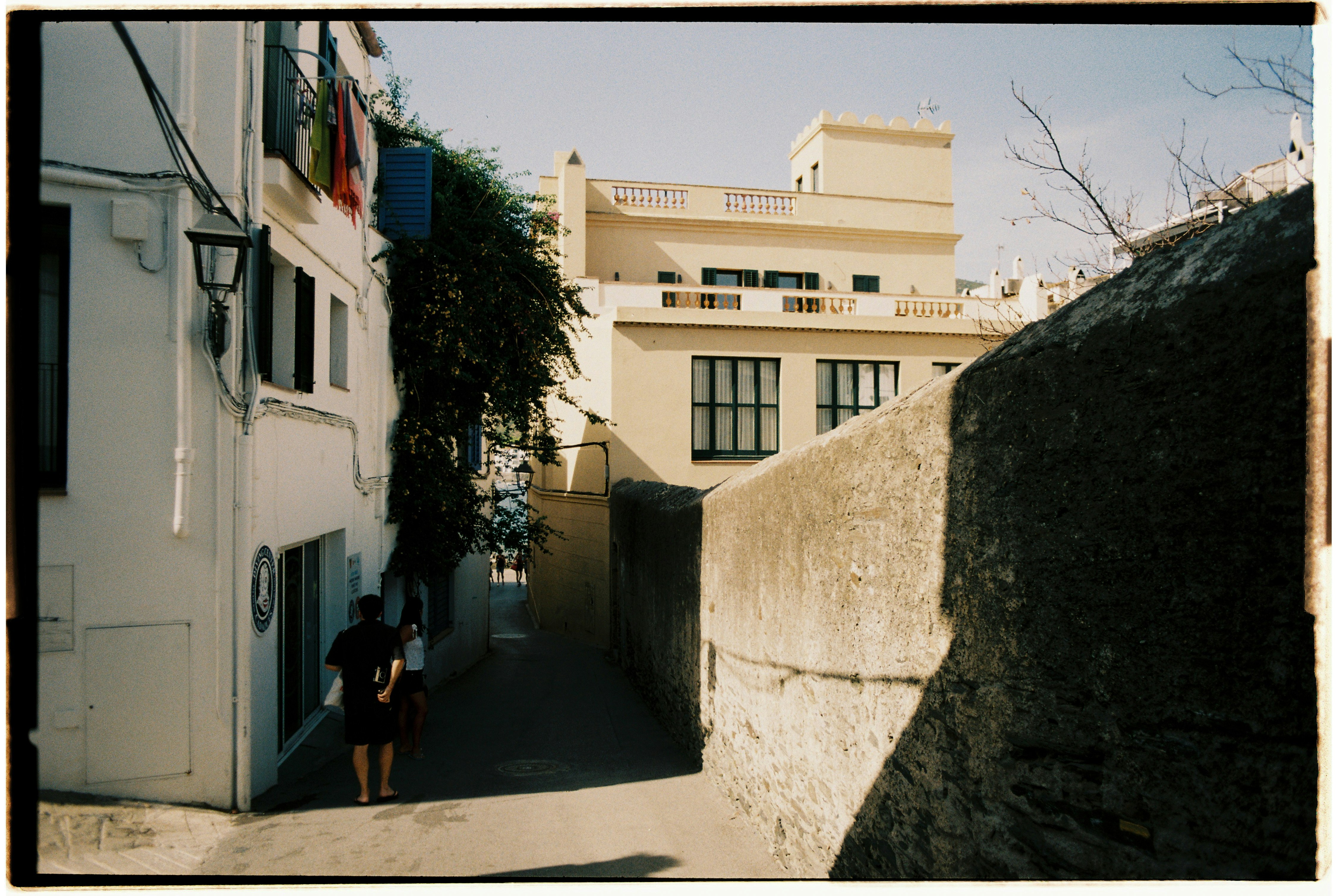 Two people walking down a narrow street between white buildings and a stone wall