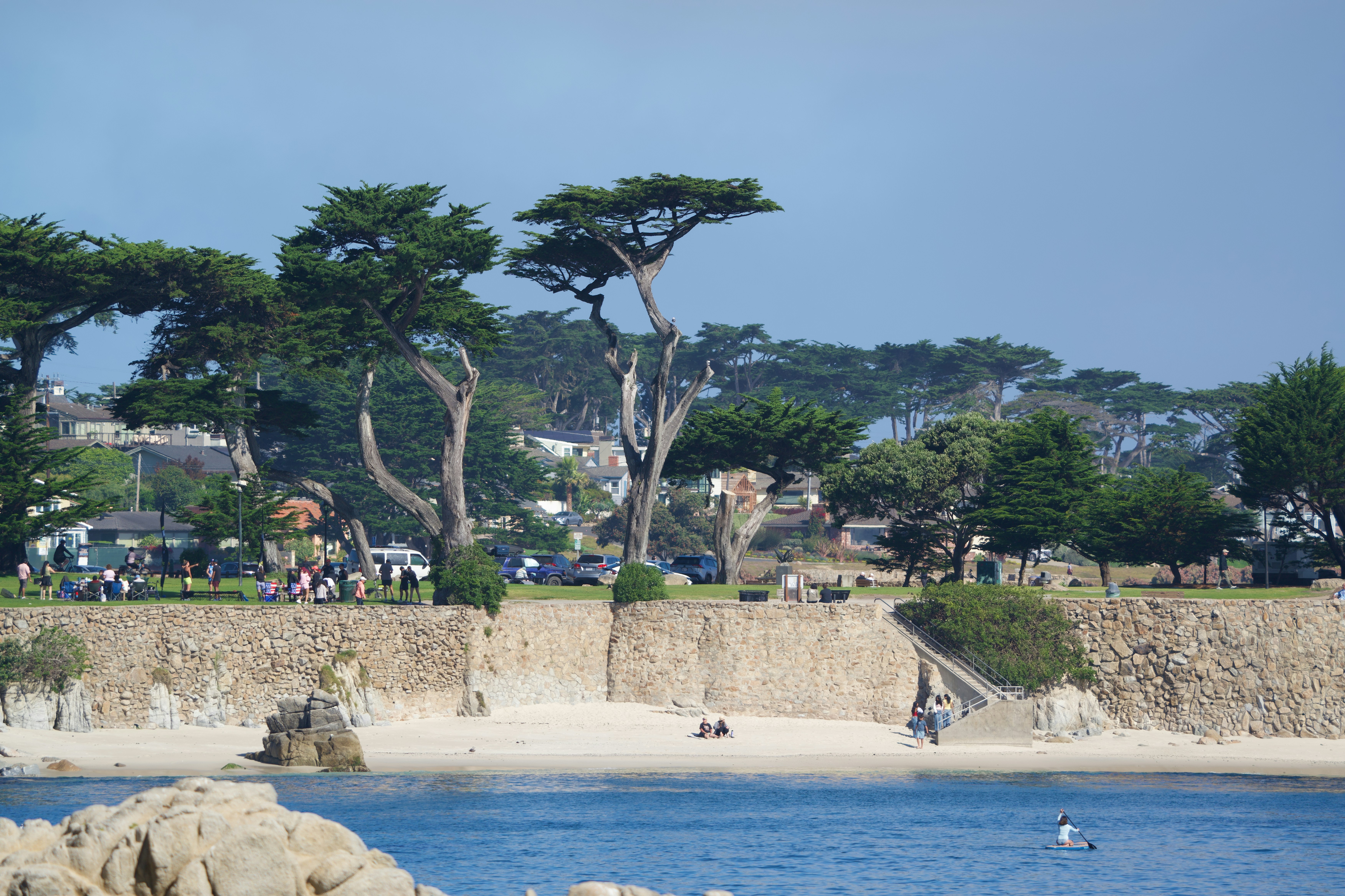 A stone wall above a sandy beach with tall cypress trees and people