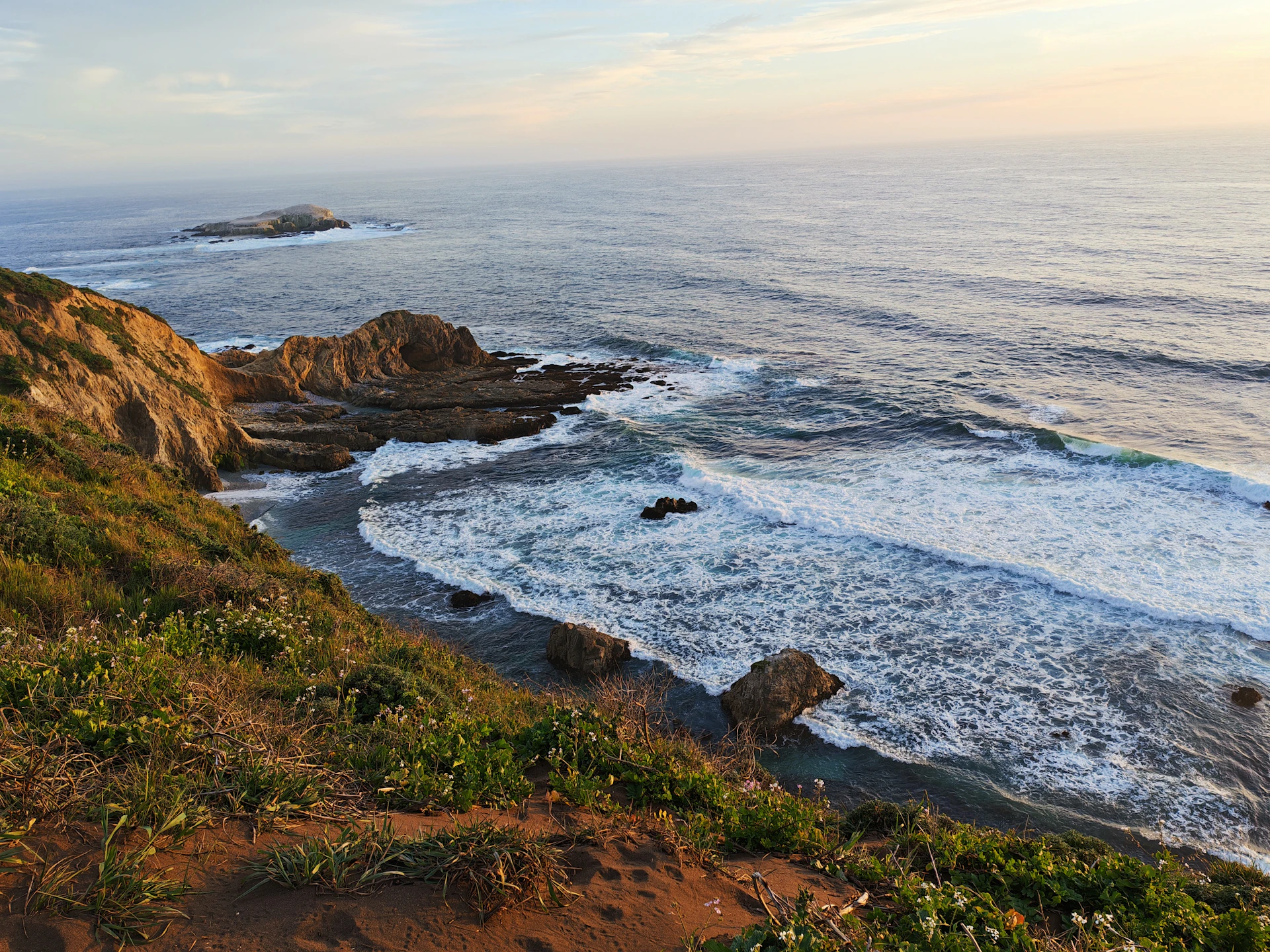 Waves crashing against rocky cliffs below a grassy hillside under a soft sky