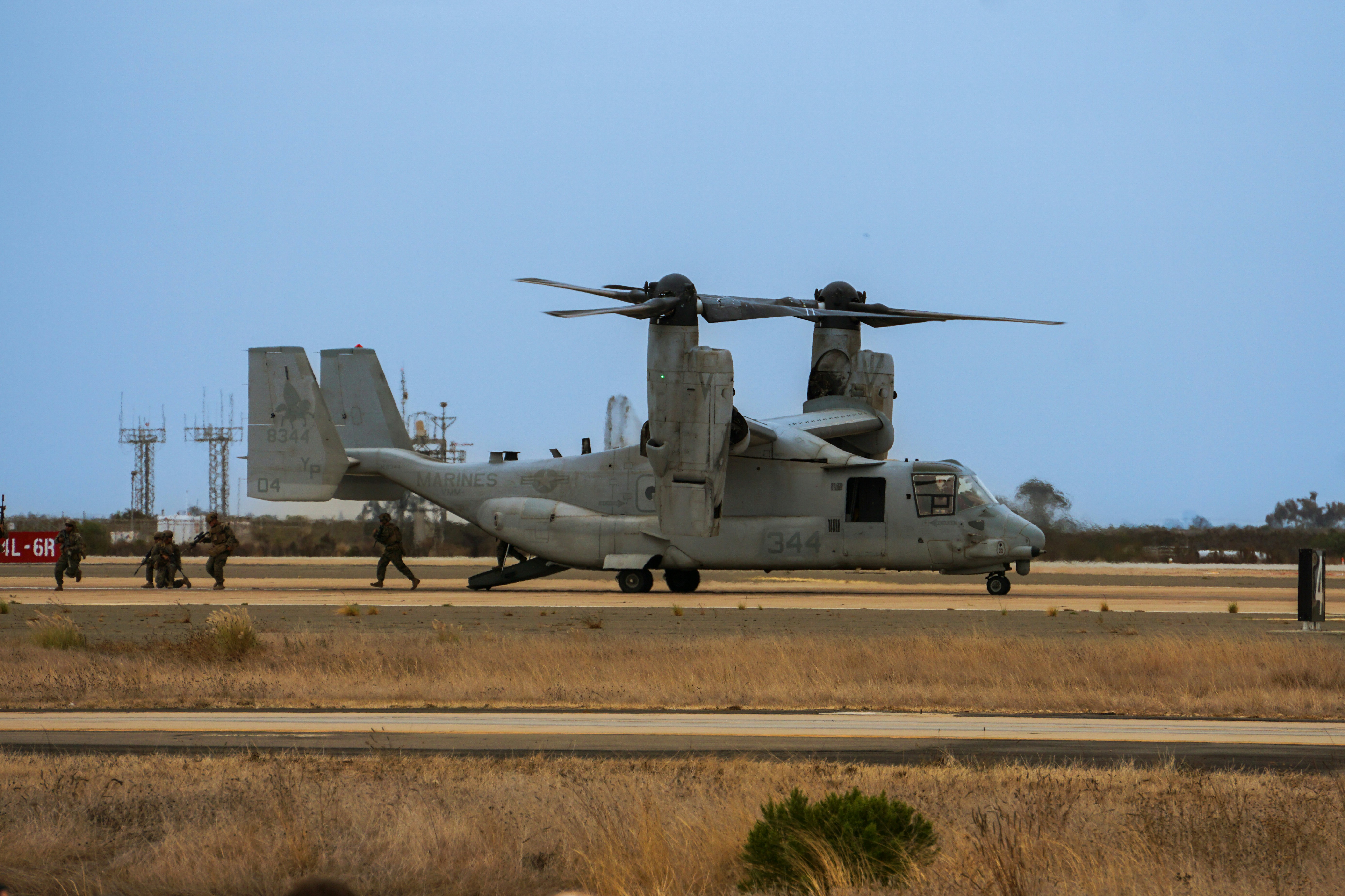 A V-22 Osprey aircraft on a runway with soldiers running nearby