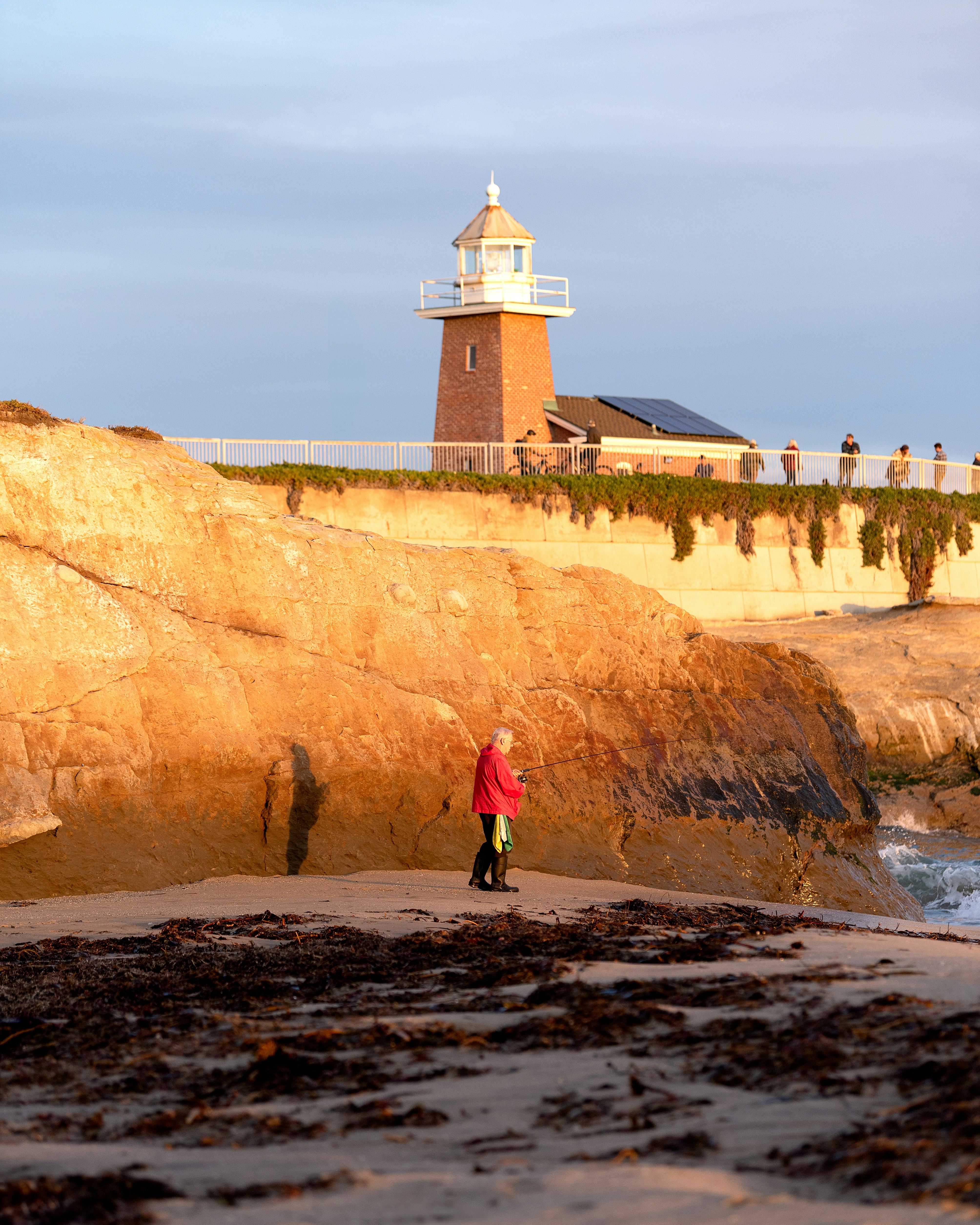 A person in a red jacket fishing on a beach below a brick lighthouse