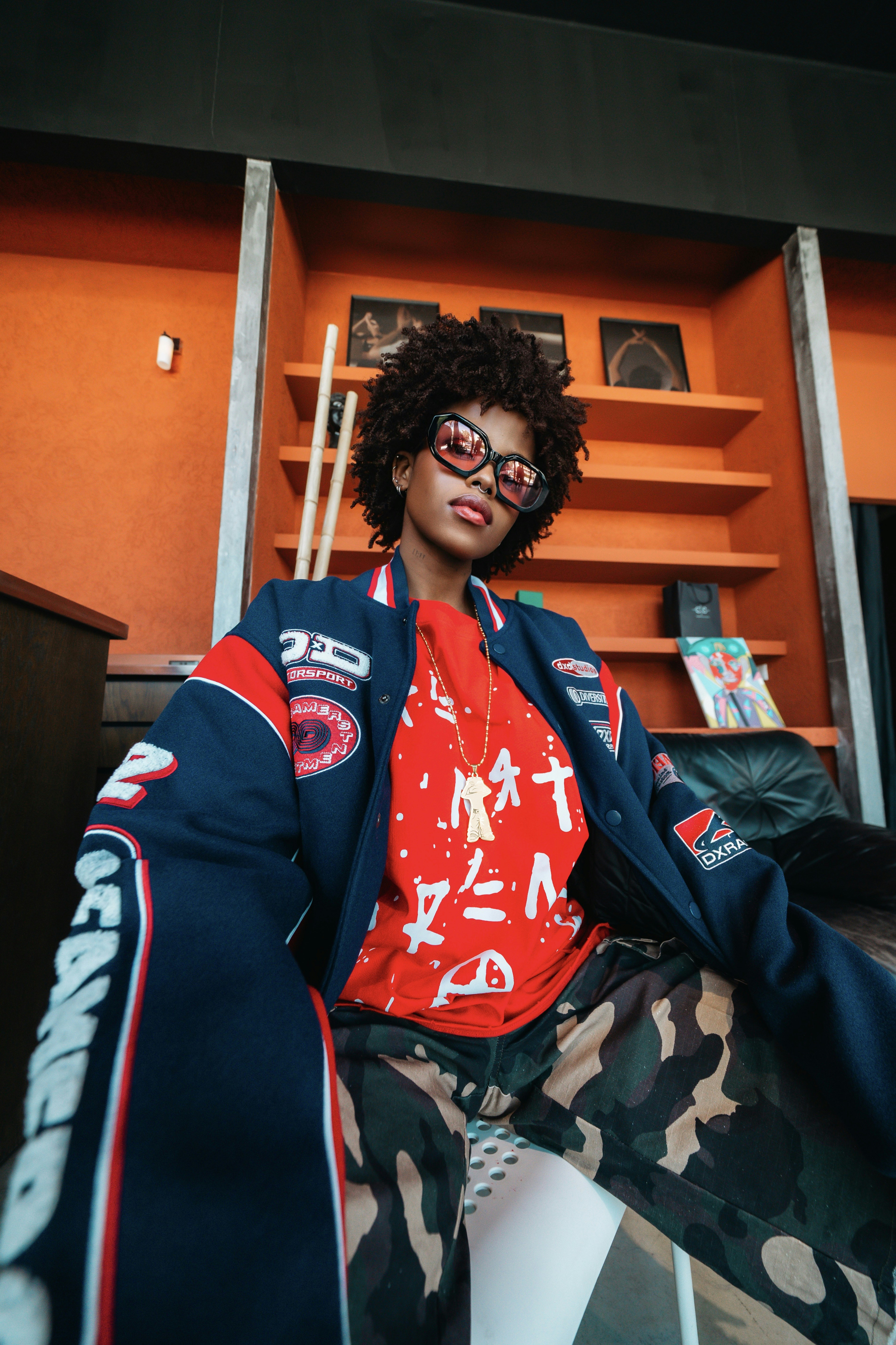 A person with curly hair wearing a varsity jacket and camouflage pants in a room