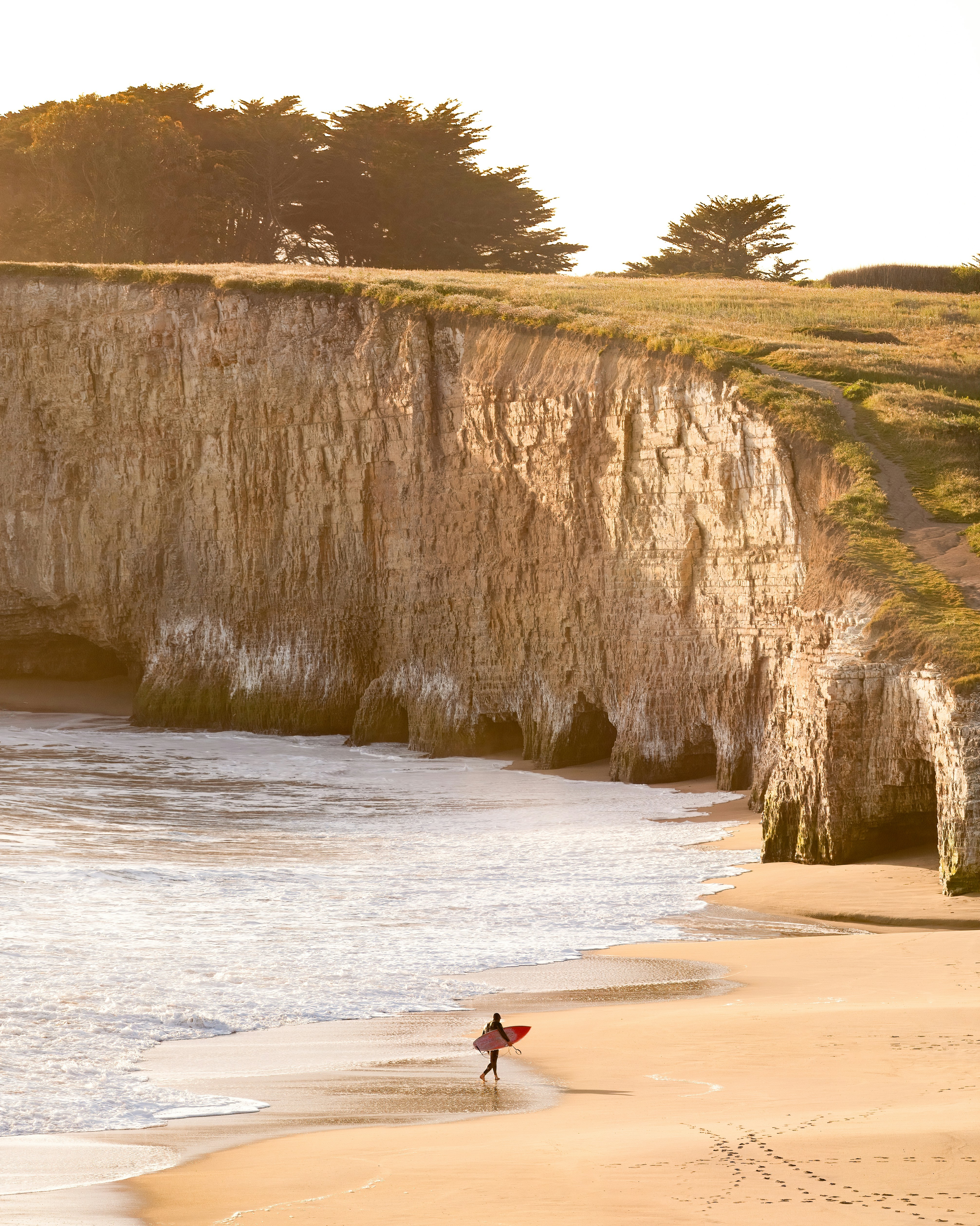 A surfer carrying a red surfboard along a sandy beach beneath tall coastal cliffs