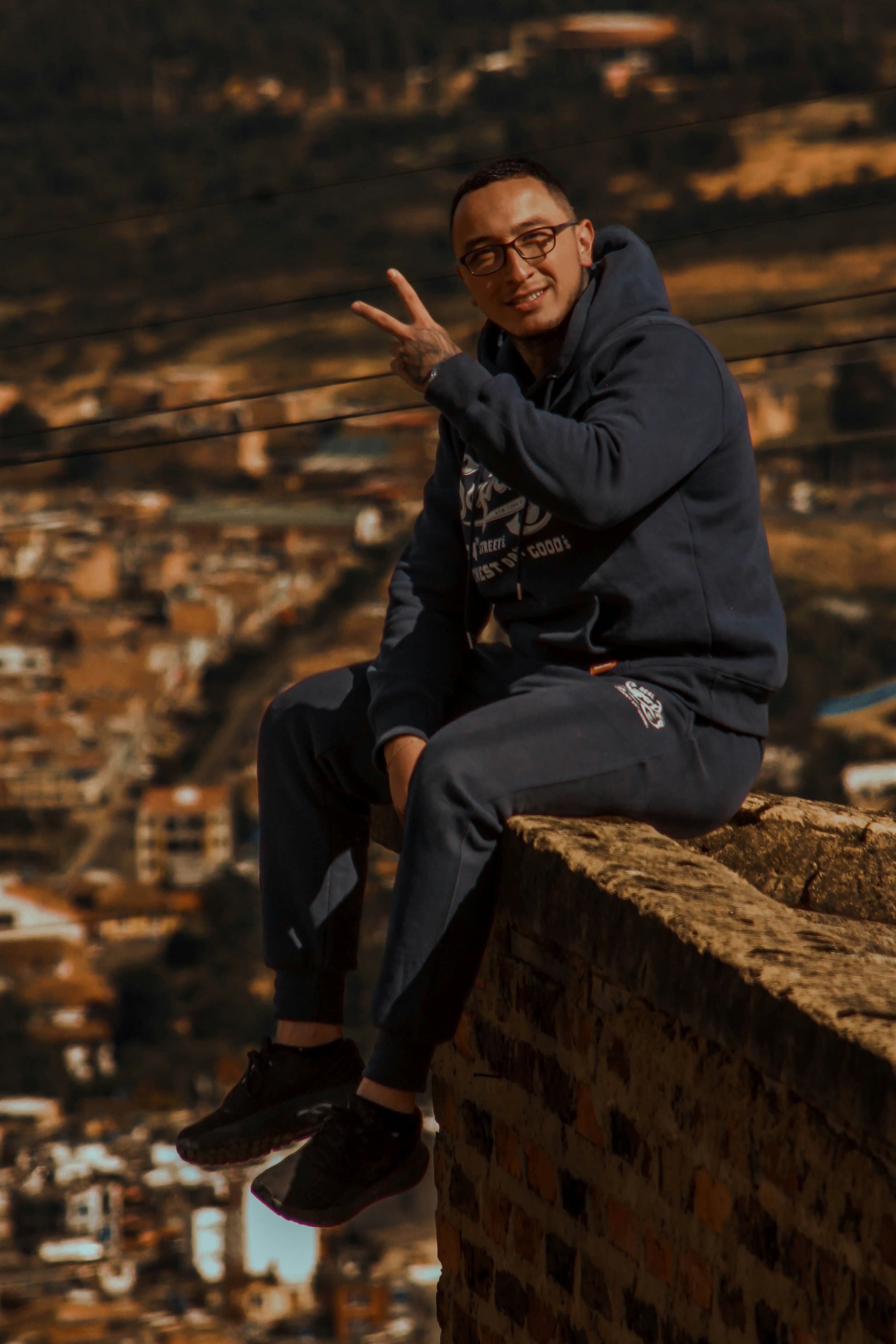 A man in a navy hoodie and sweatpants making a peace sign on a wall