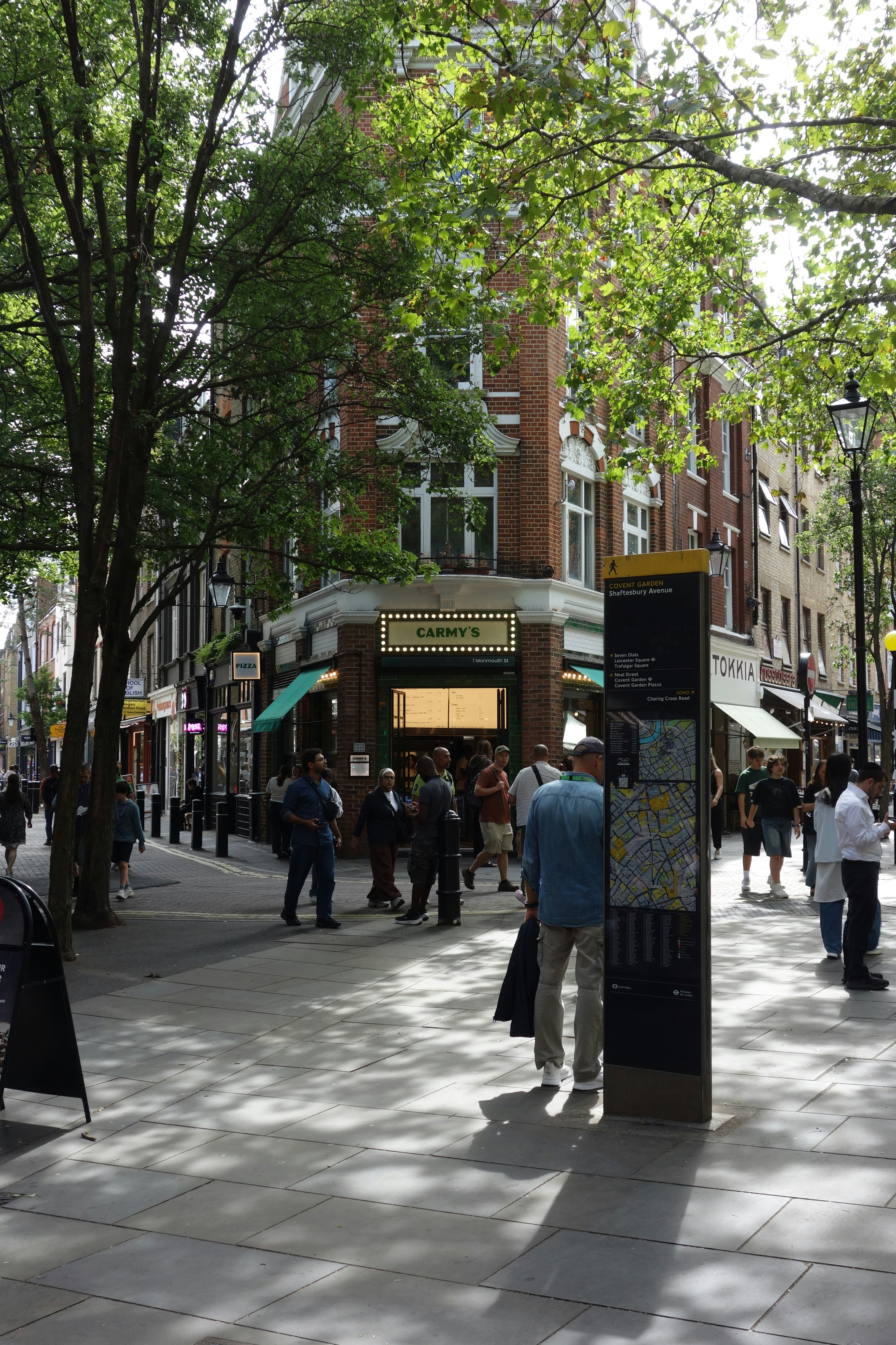 People walking on a sunlit street in London near a wayfinding map sign