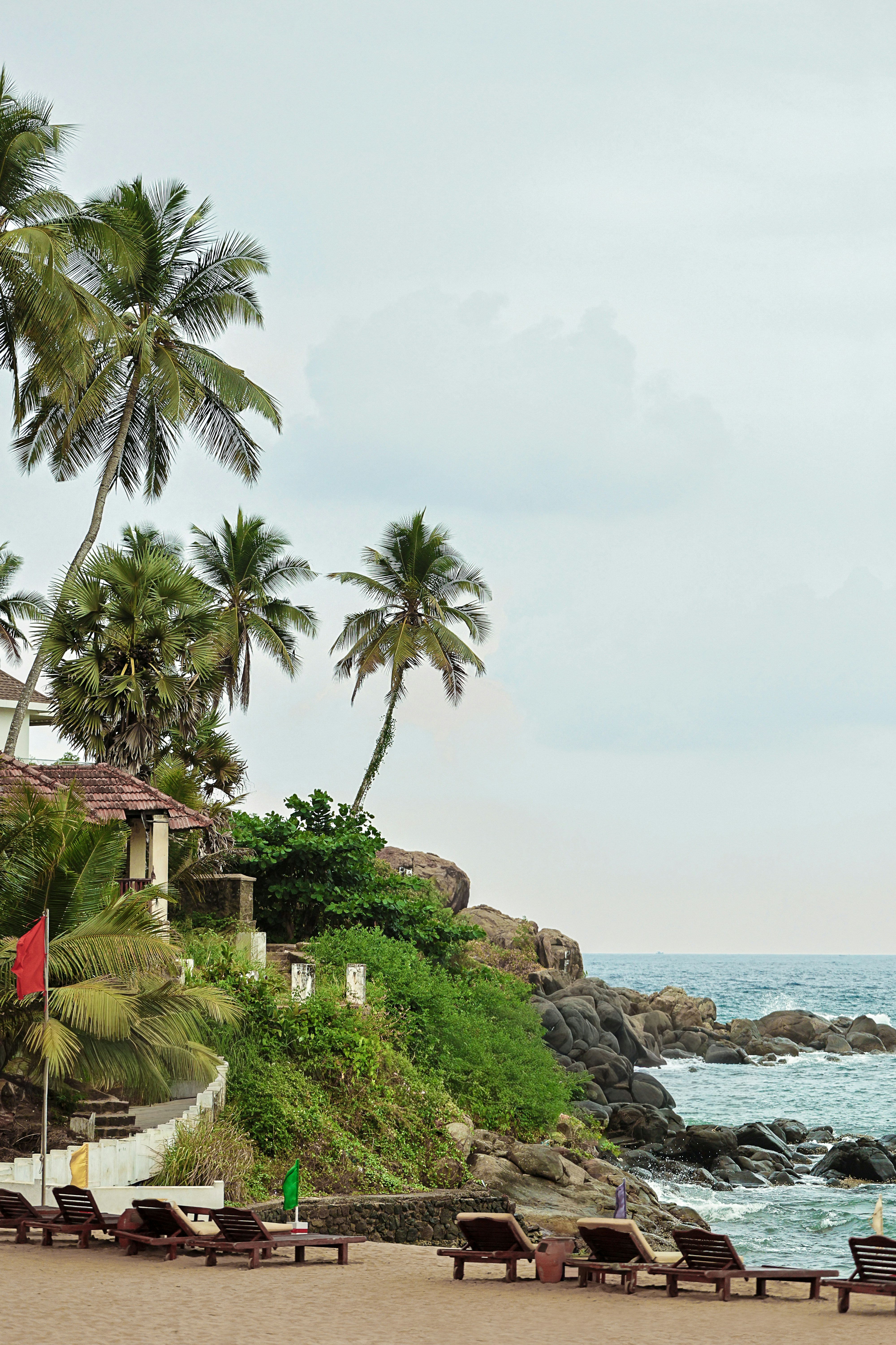 Wooden lounge chairs on a sandy beach below palm trees and rocky cliffs