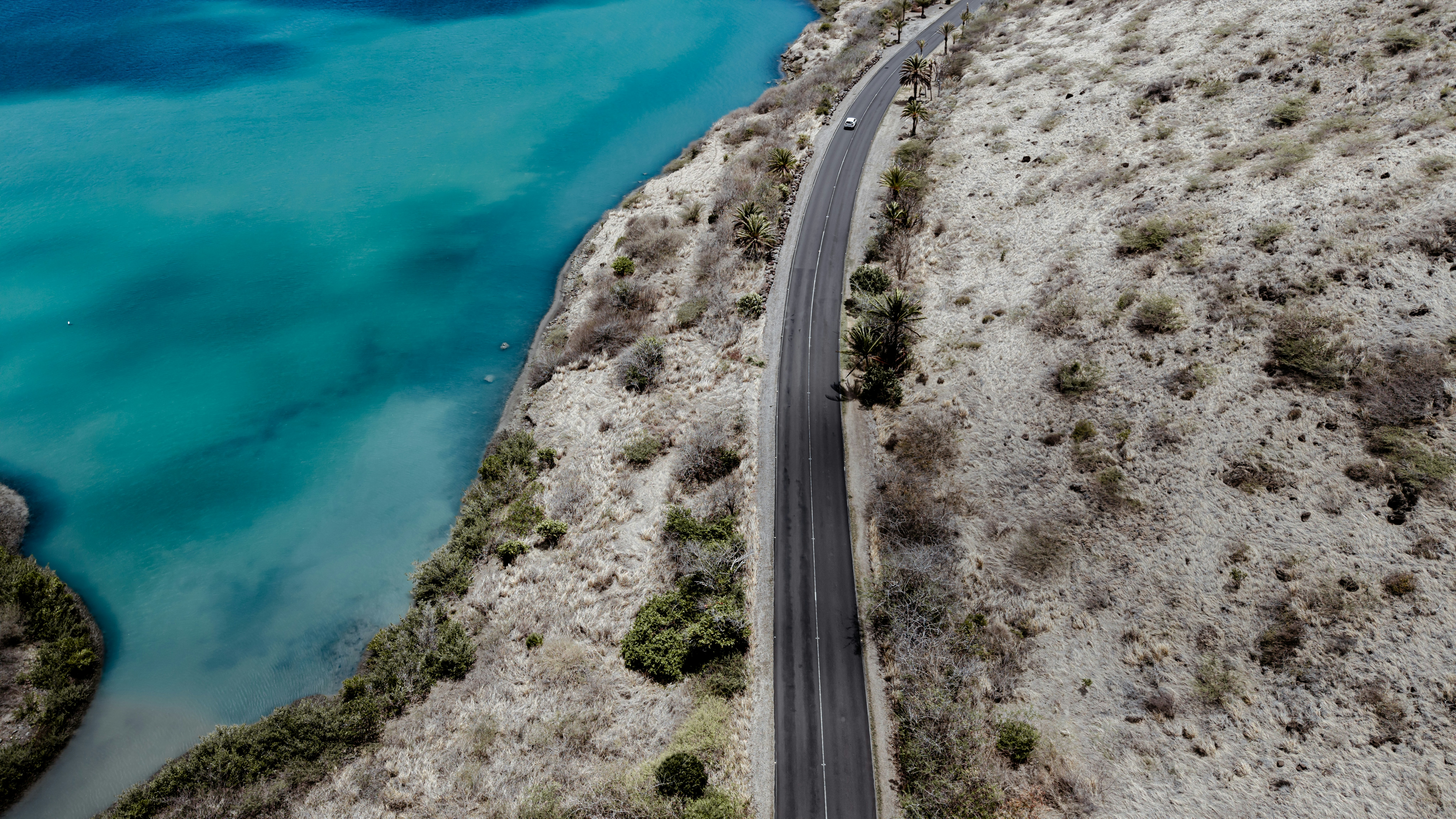 An aerial view of a coastal road beside turquoise water and dry hills