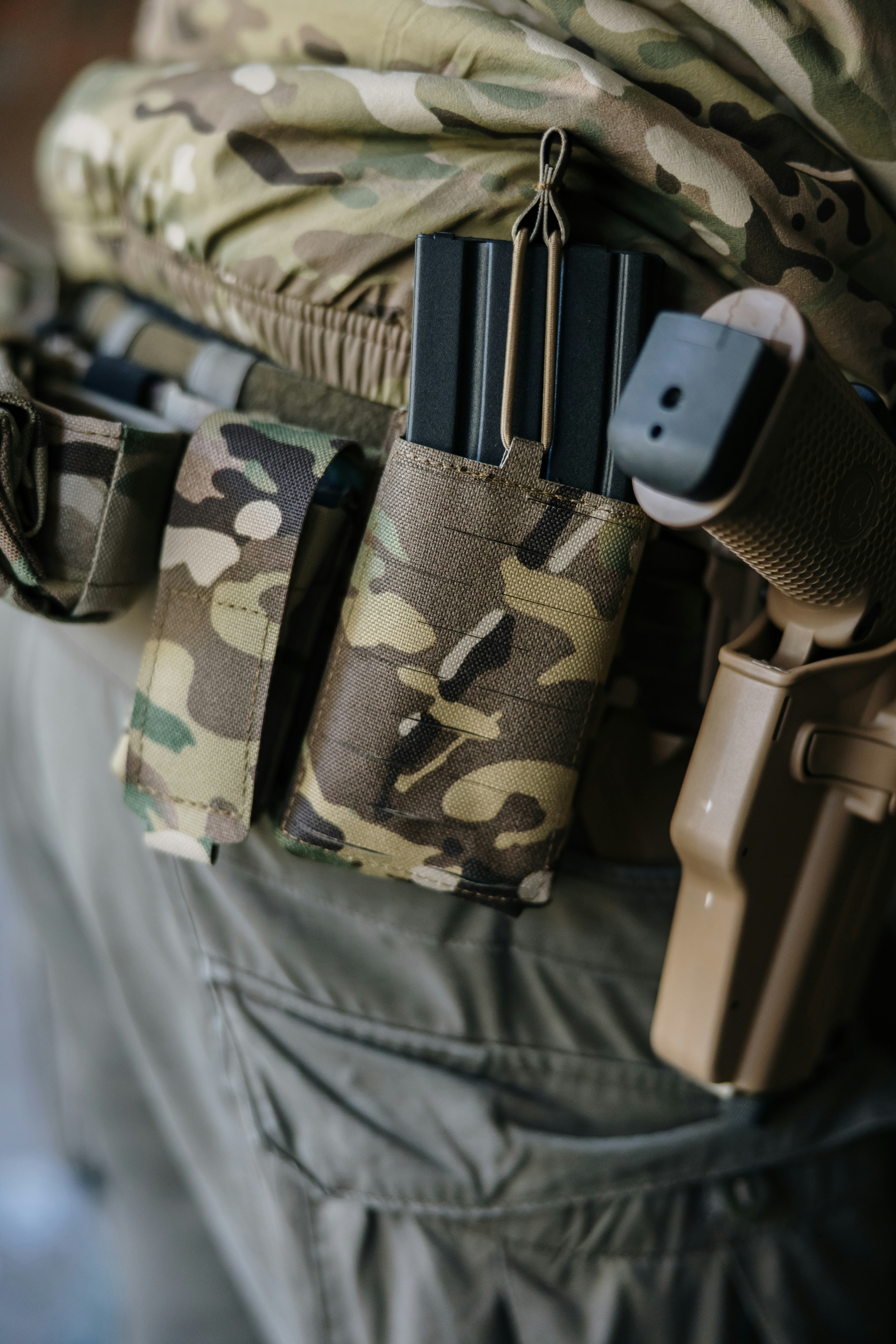 A black airsoft magazine held in a camouflage pouch beside a tan holster, captured in a close-up of the player’s tactical belt.