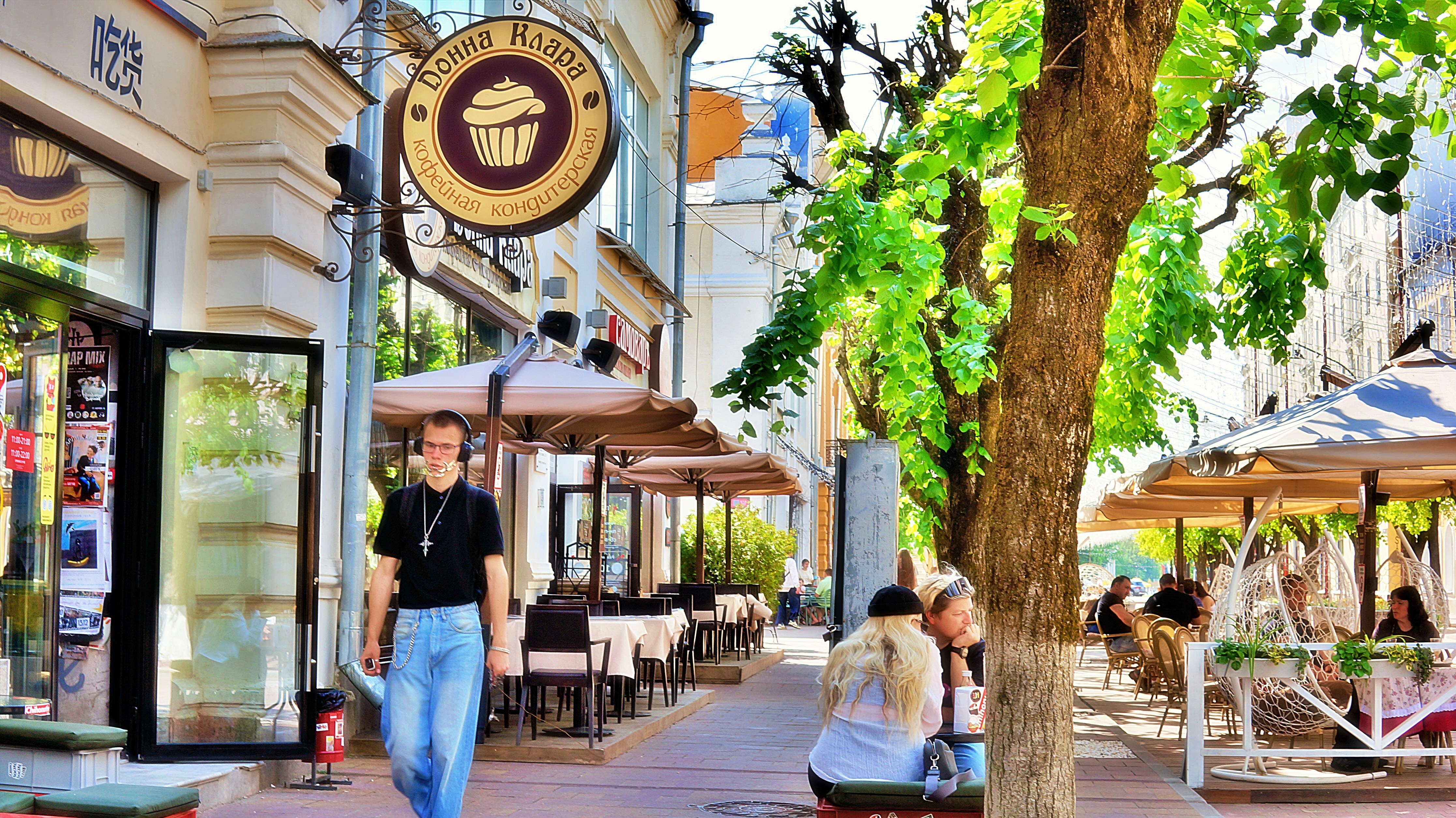 A person walking past outdoor cafe tables on a sunny tree-lined city street