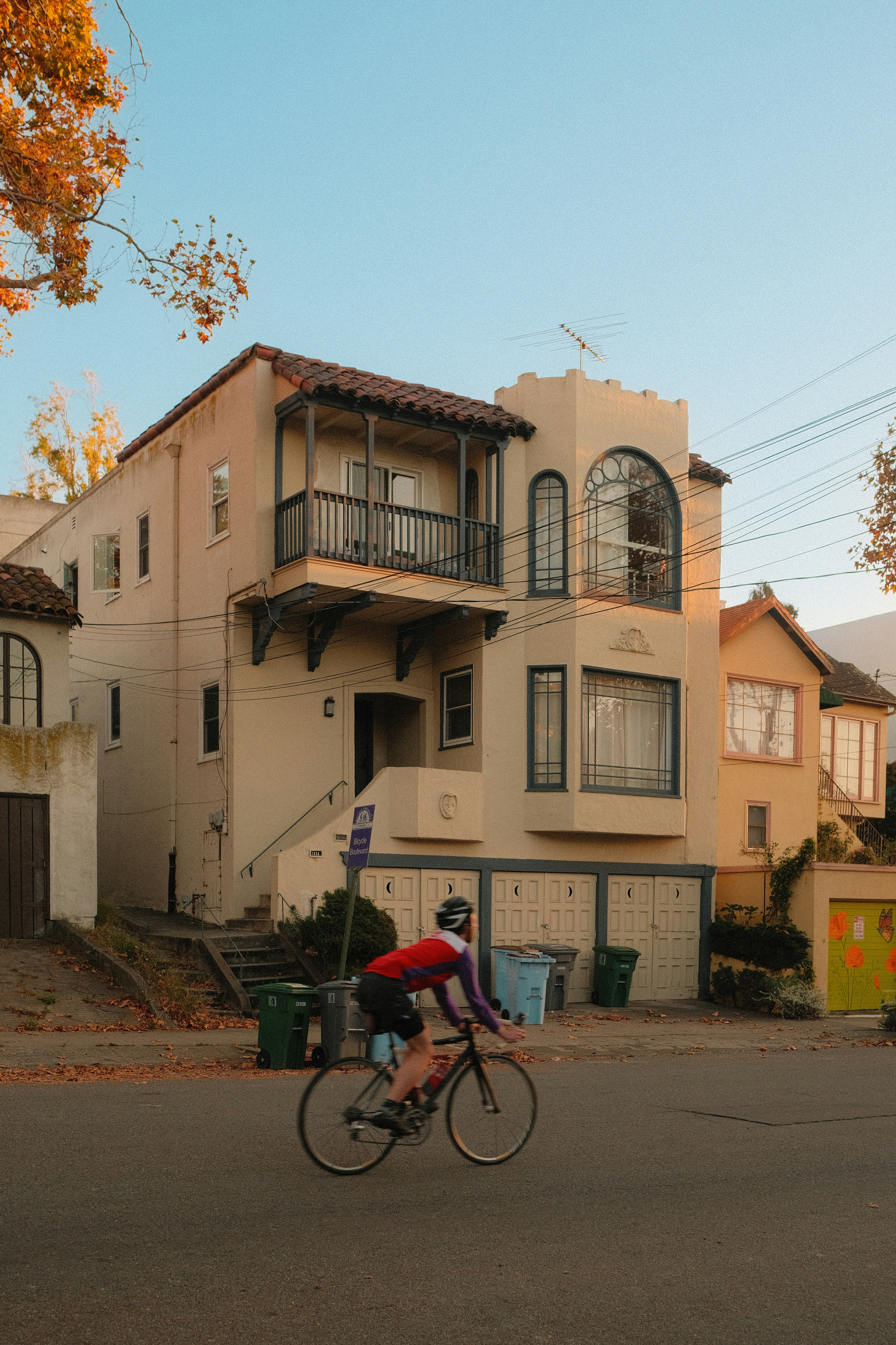 A person cycling past a beige stucco house with a balcony and arched windows