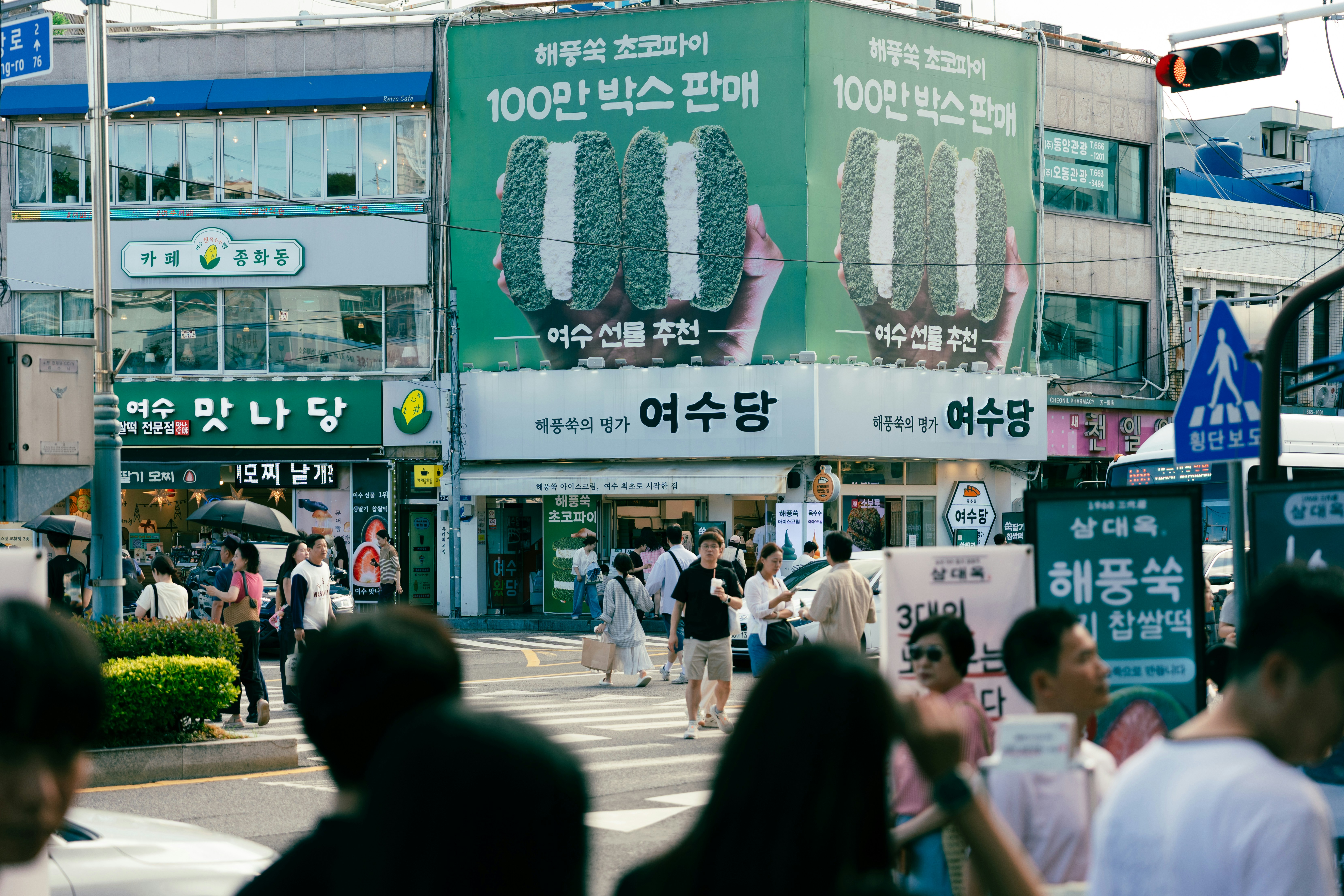People walking across a busy street intersection in Yeosu with large green advertisements