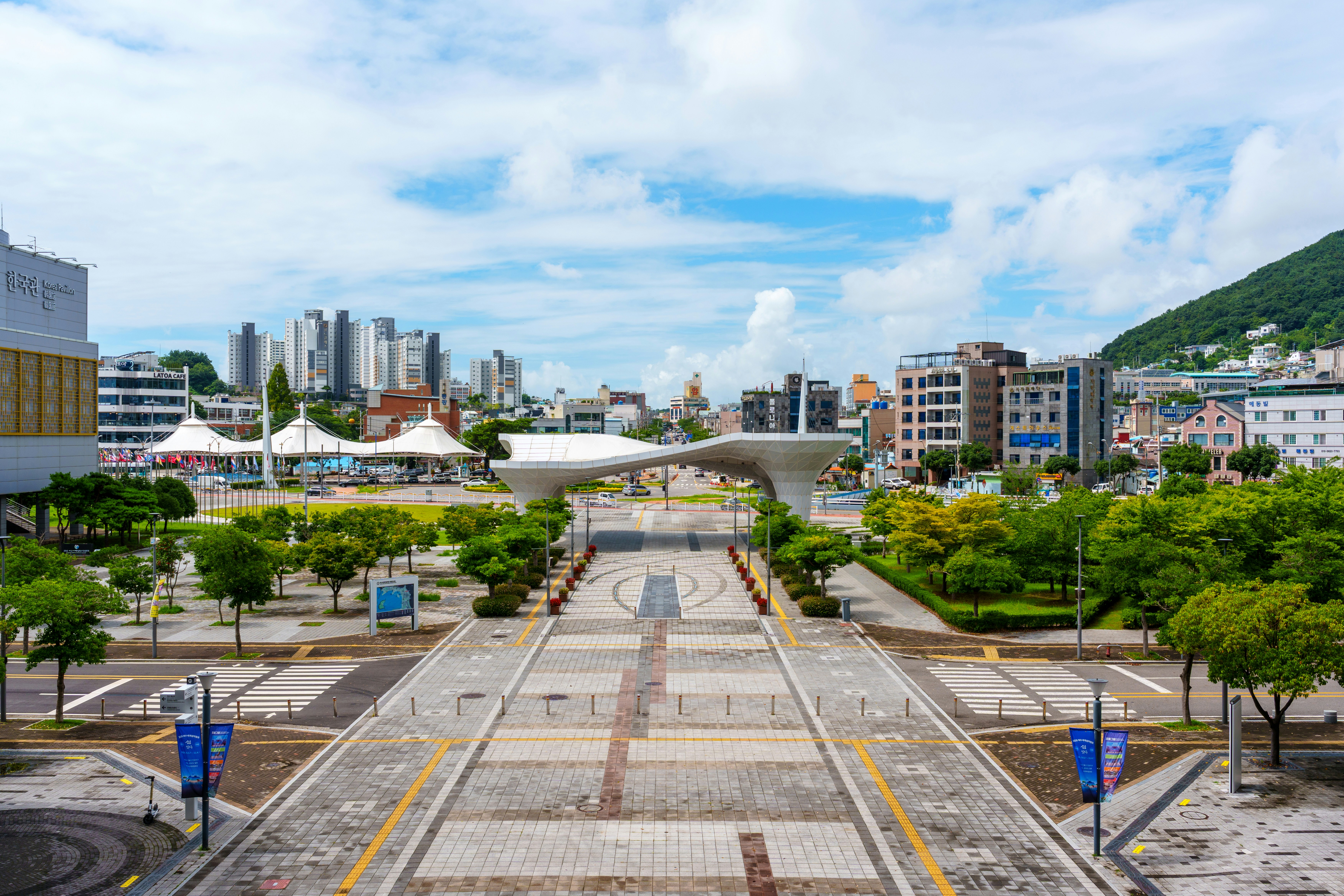 A paved plaza with a modern white canopy structure leading toward a city skyline
