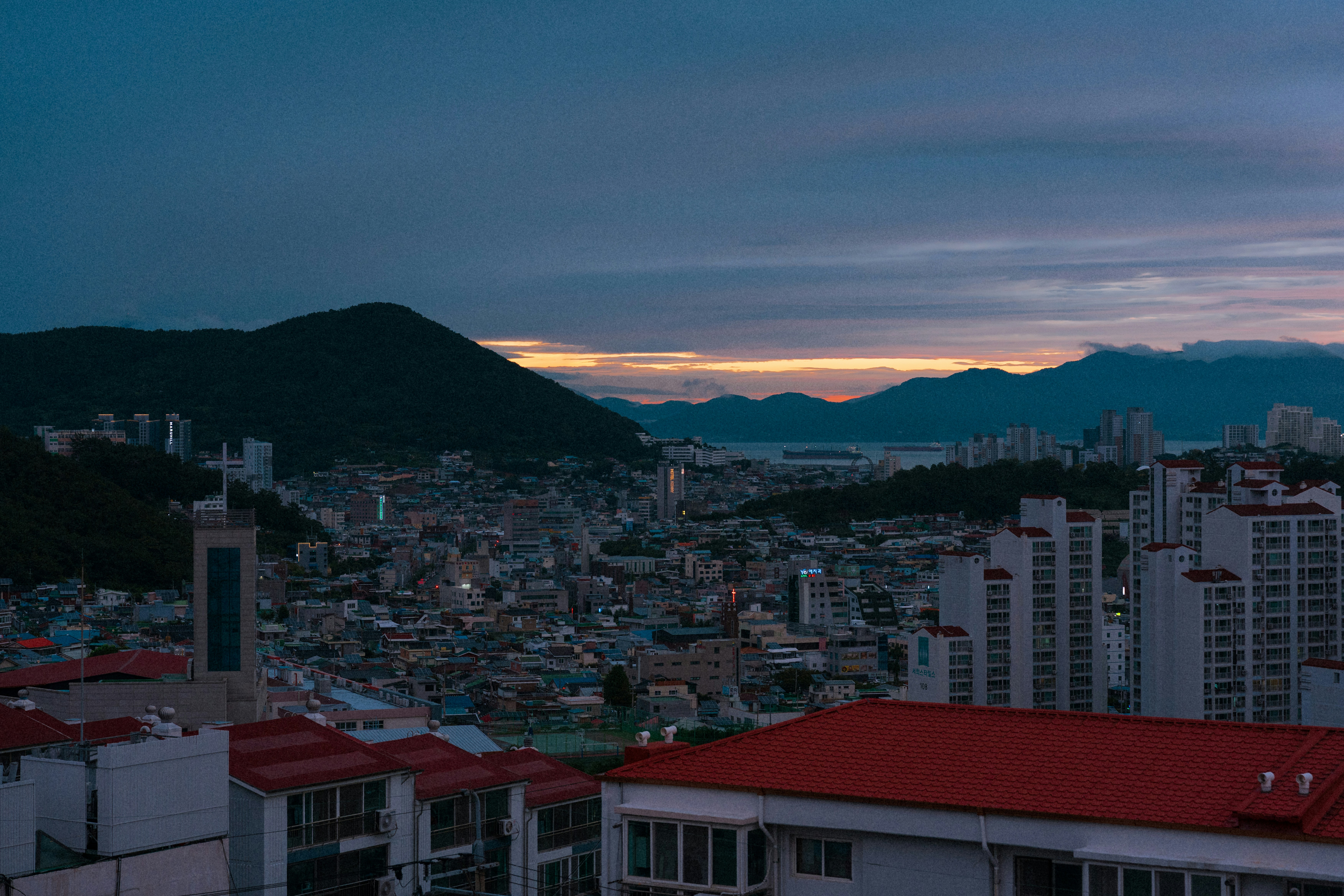 A dense city with red-roofed buildings nestled between mountains under a dark sky
