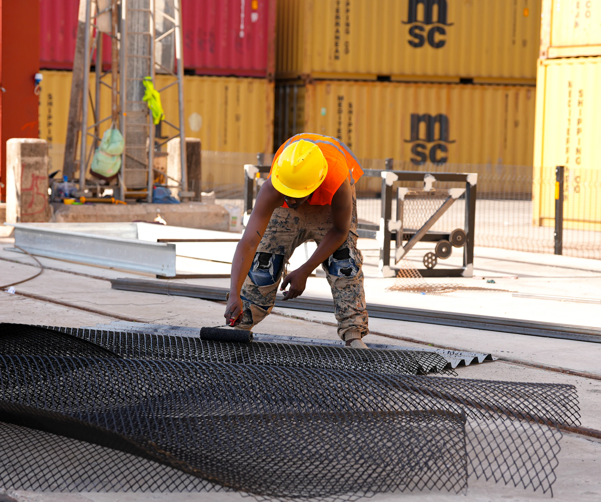 A construction worker in a yellow hard hat and orange vest painting black mesh on a concrete floor