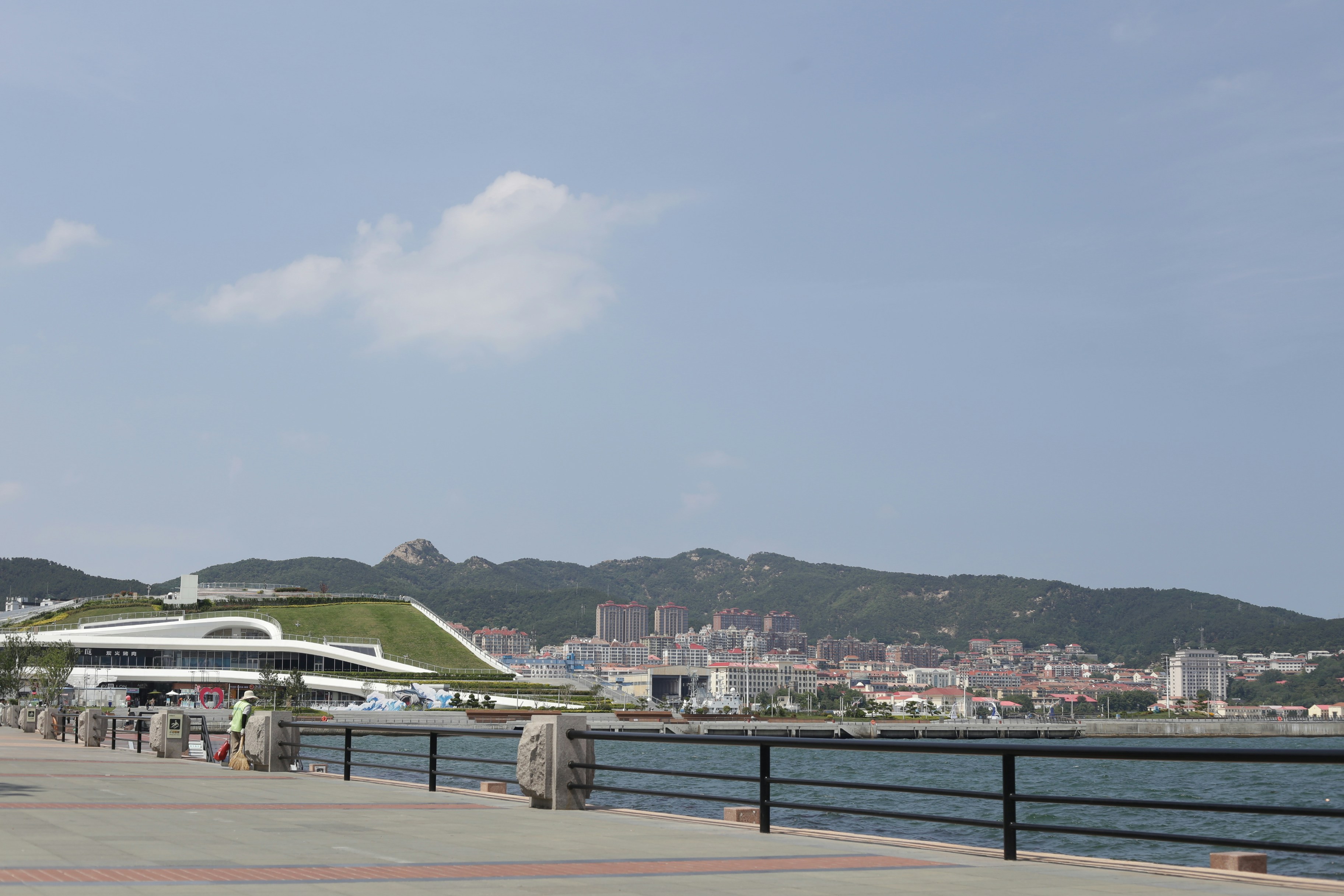A paved waterfront promenade with a modern building and distant mountains under a blue sky