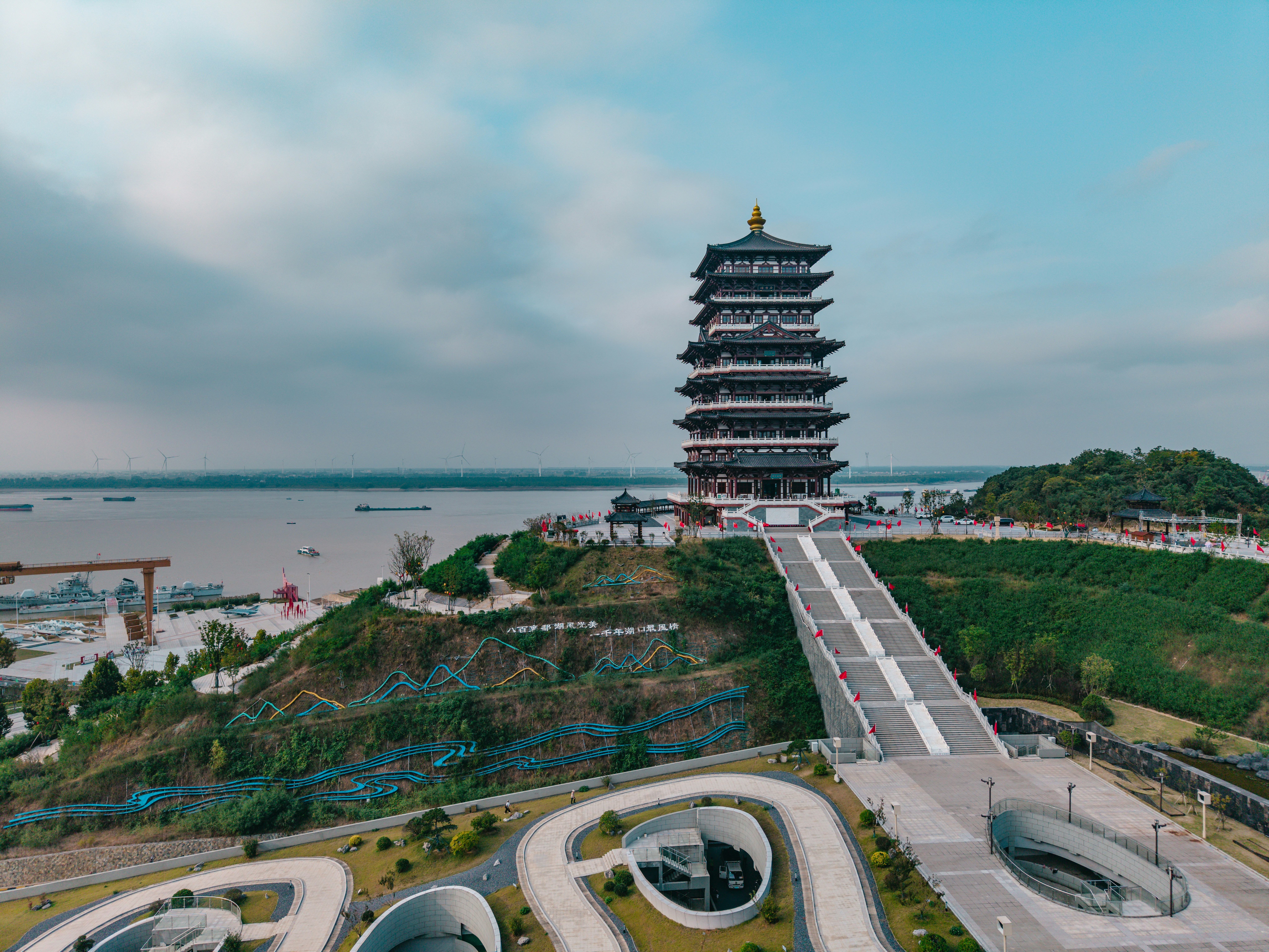 A tall pagoda on a grassy hill overlooking a wide river with wind turbines