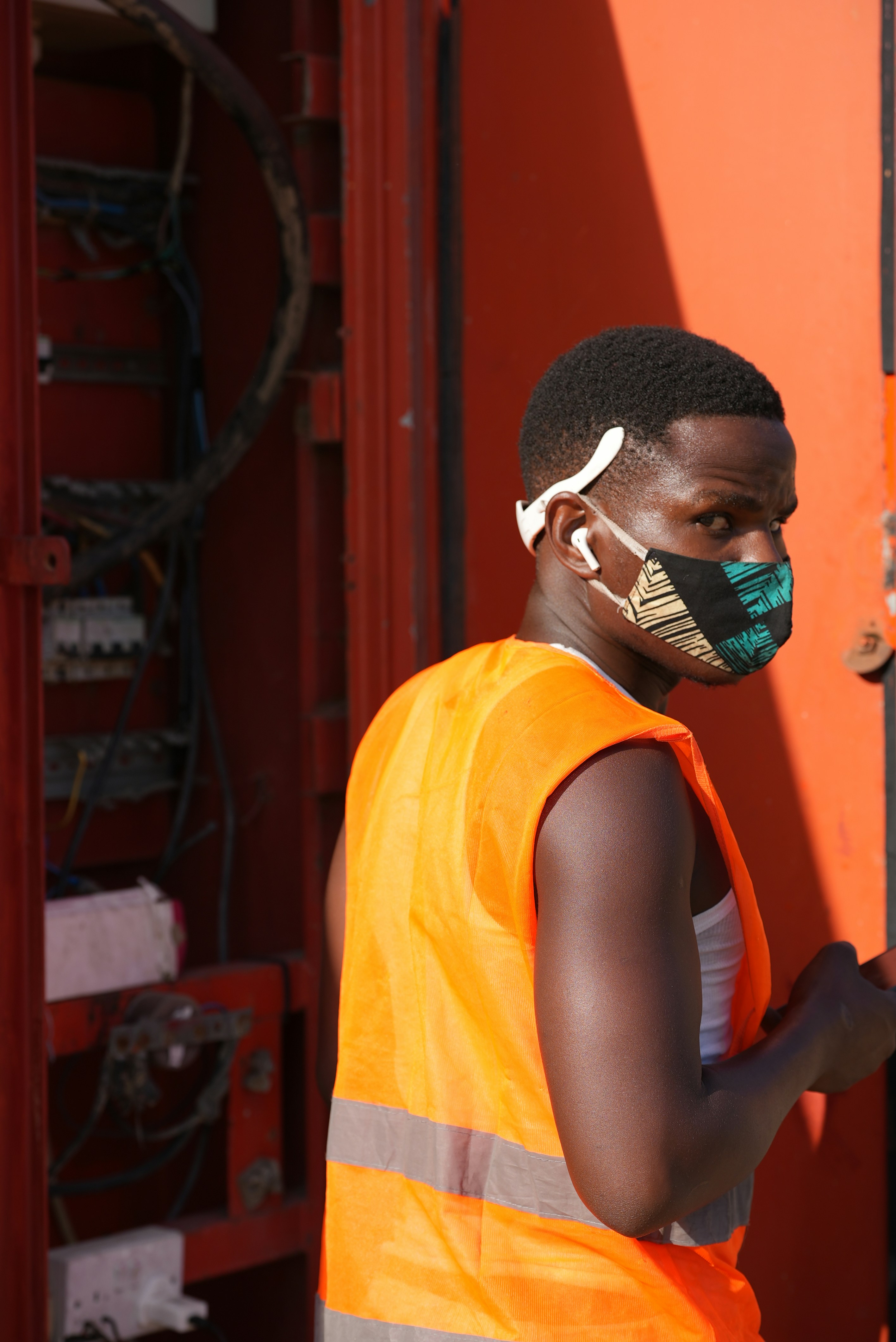A man in an orange safety vest and patterned face mask looking back at an electrical panel