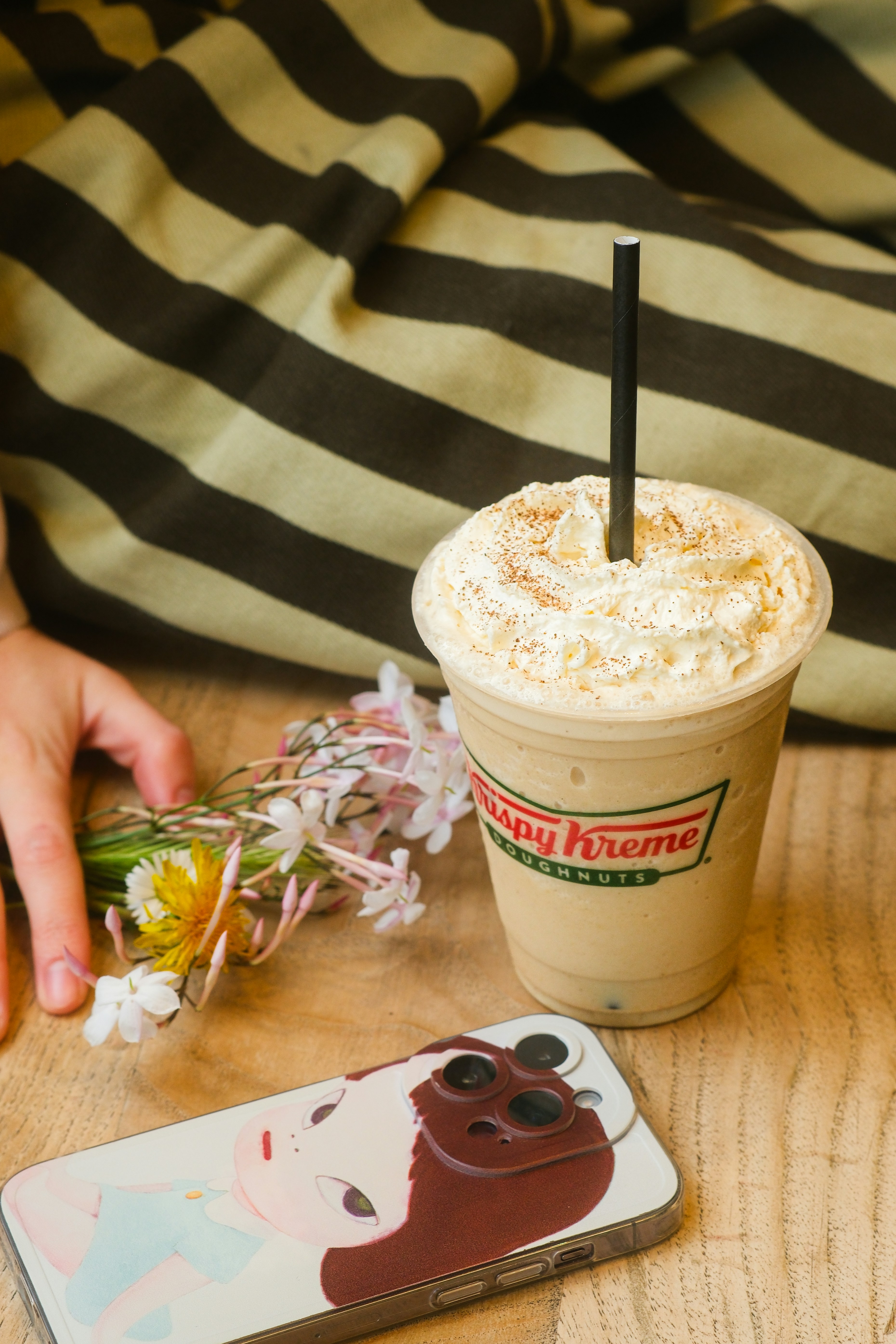A Krispy Kreme iced coffee with whipped cream beside flowers and a phone