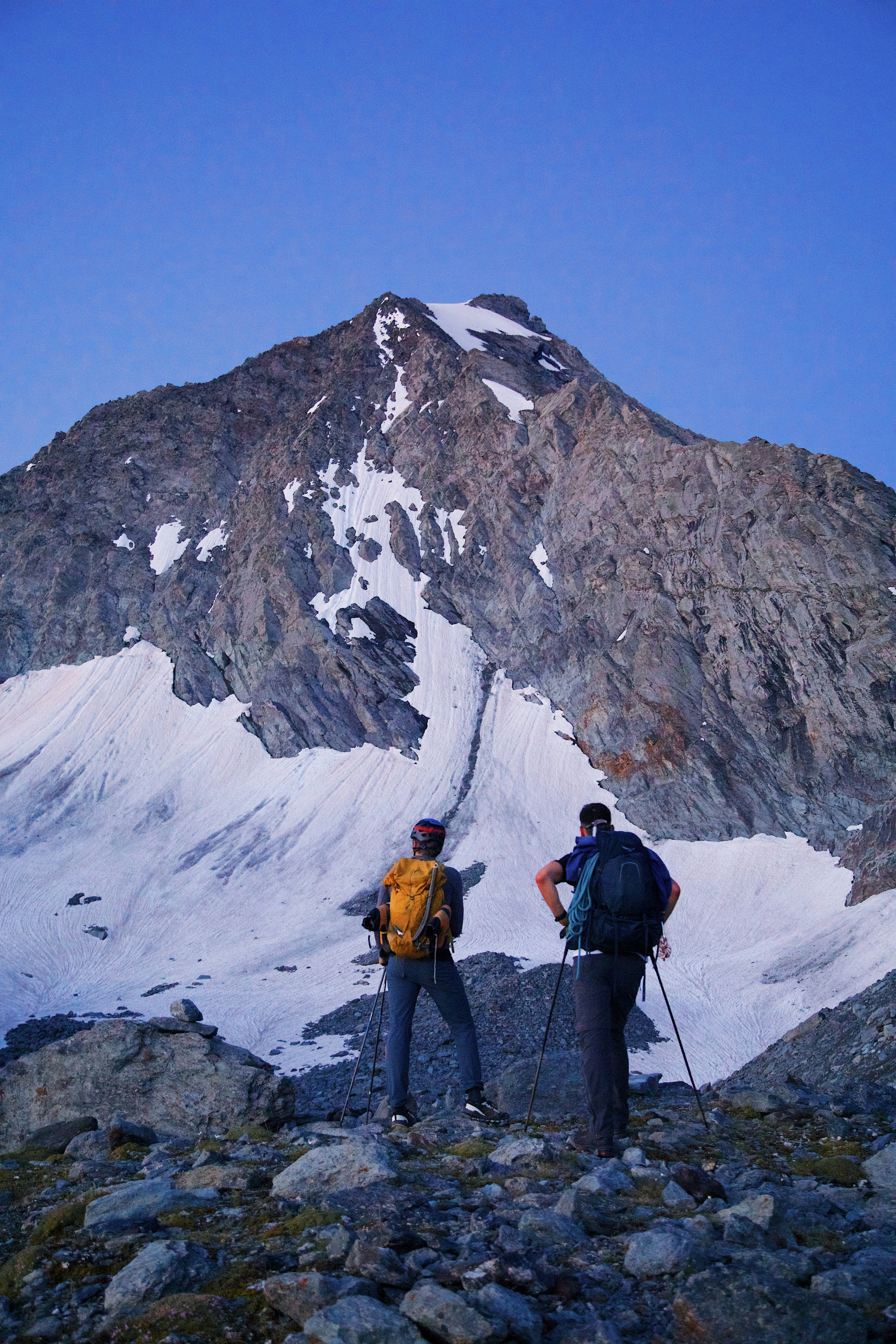 Two hikers with backpacks and trekking poles facing a steep snow-covered mountain peak