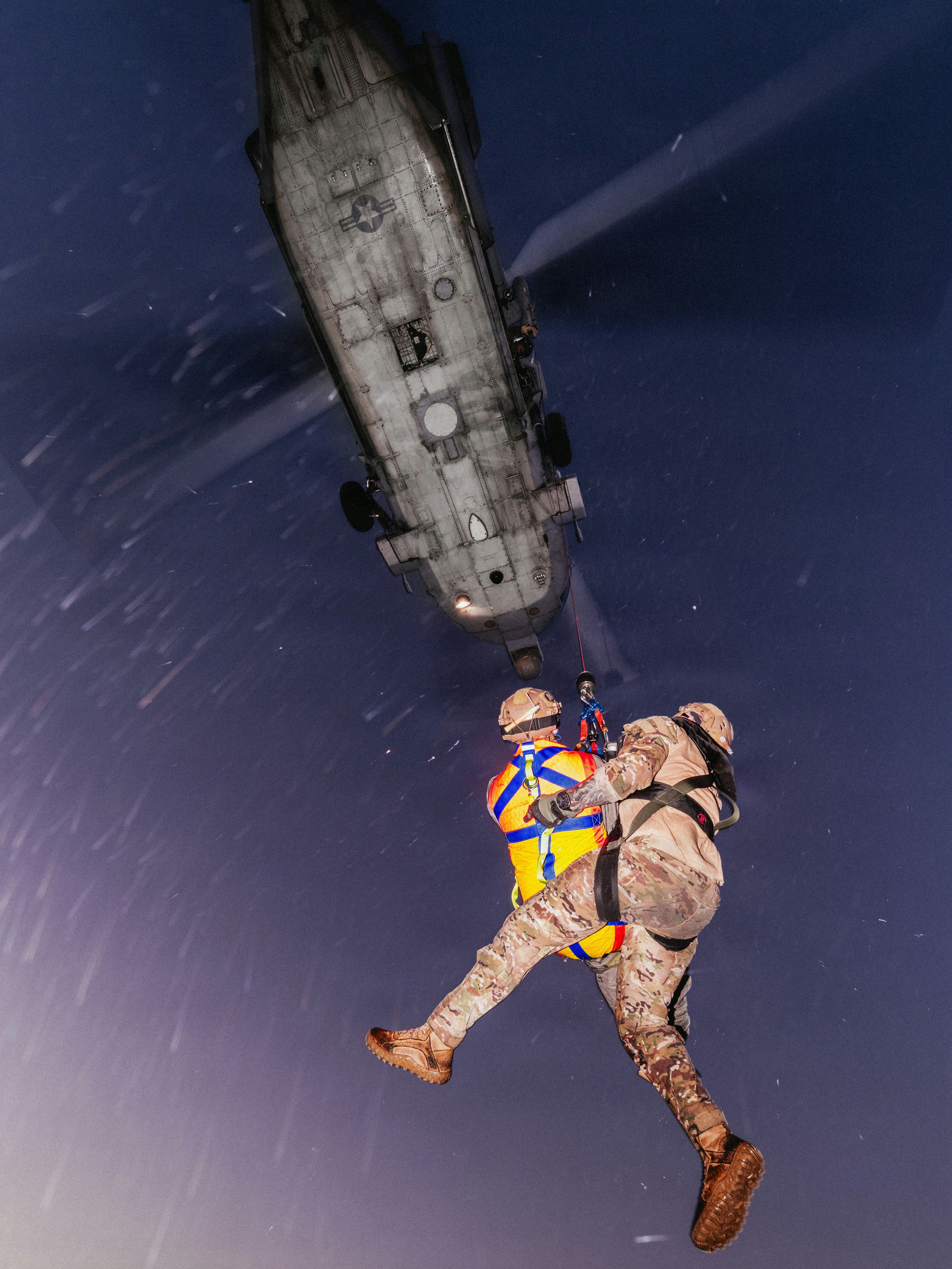 A military helicopter hoisting two people in harnesses during a night rescue operation