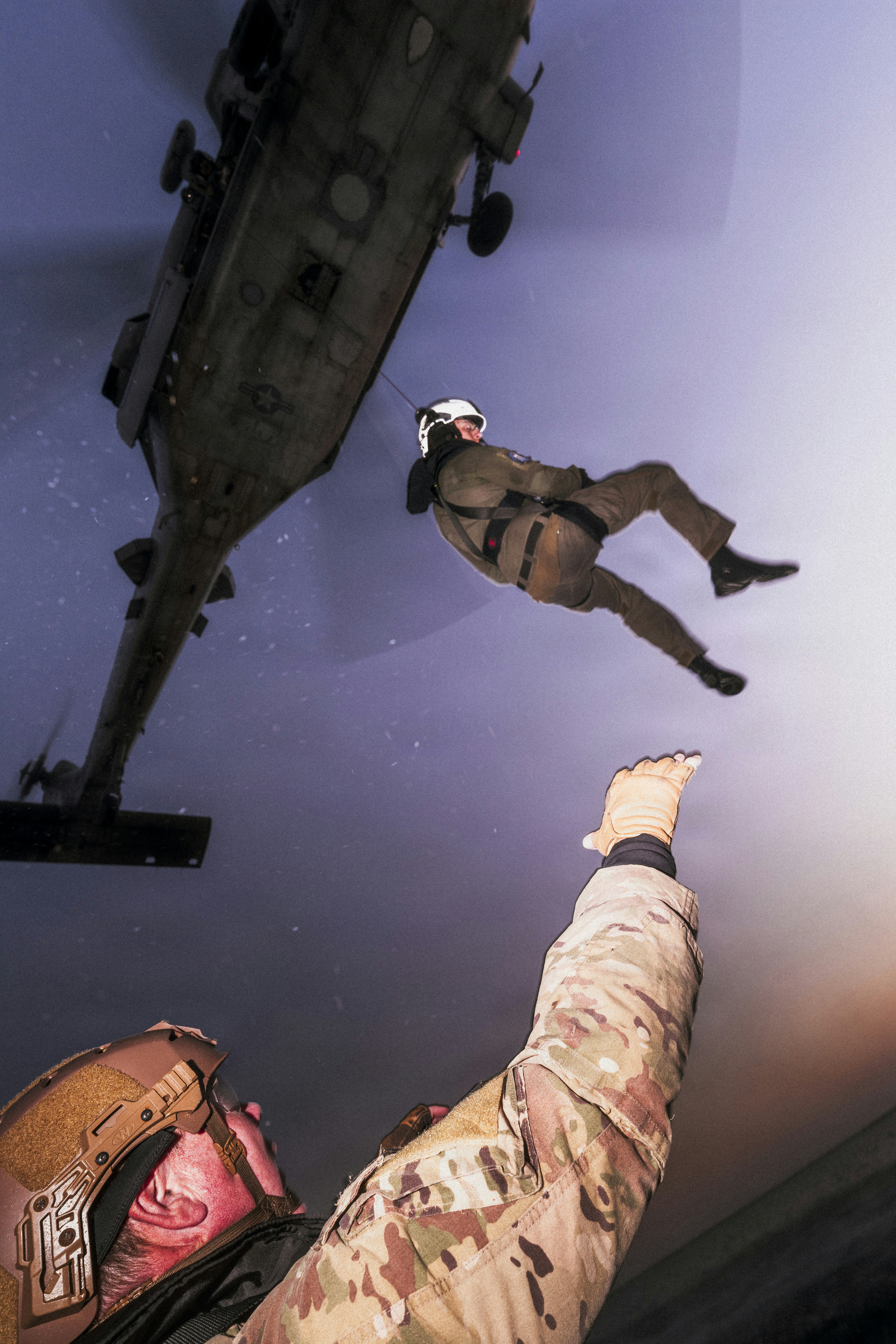A person in a harness descending from a military helicopter toward a soldier