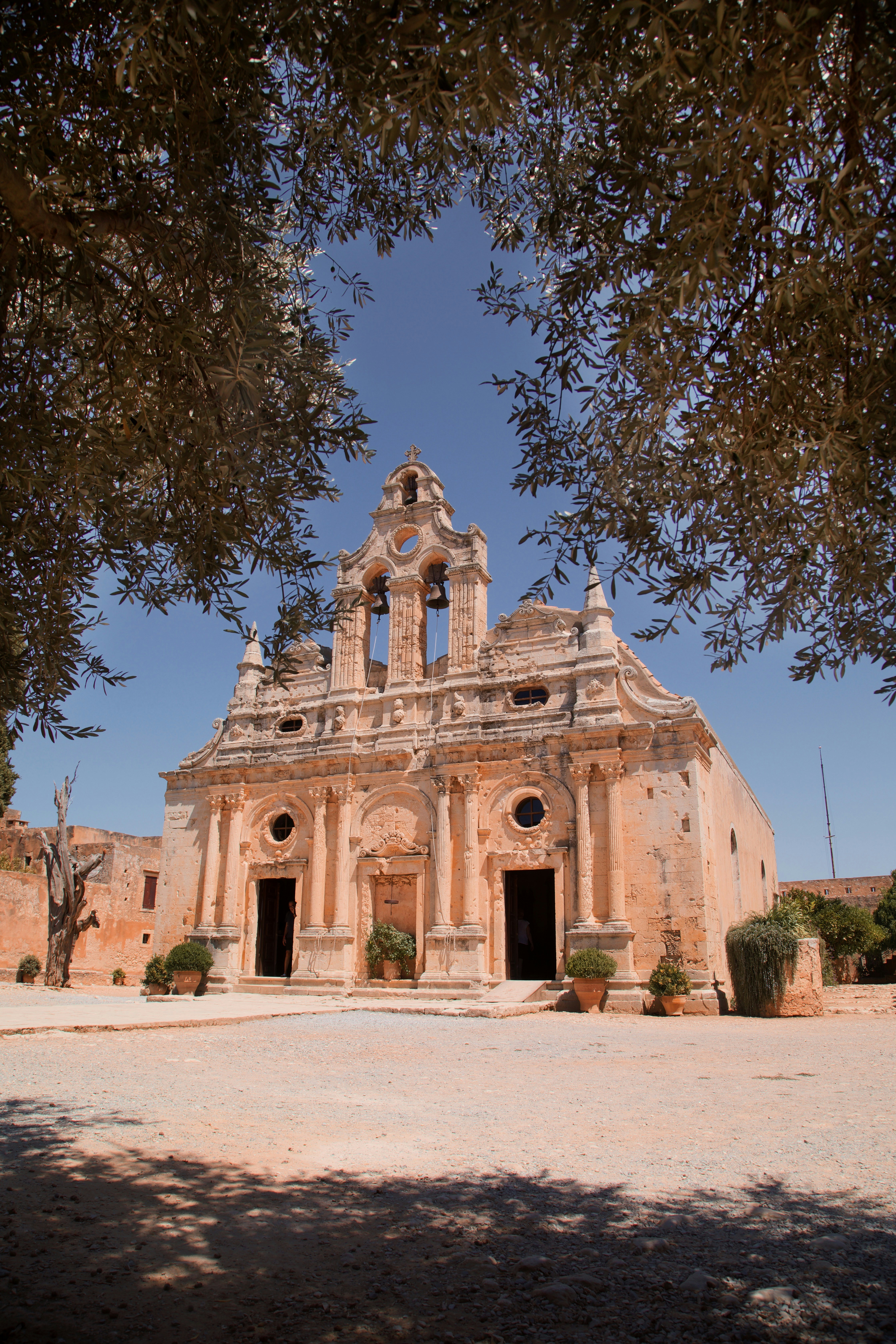 The stone facade of Arkadi Monastery framed by olive tree branches under a blue sky