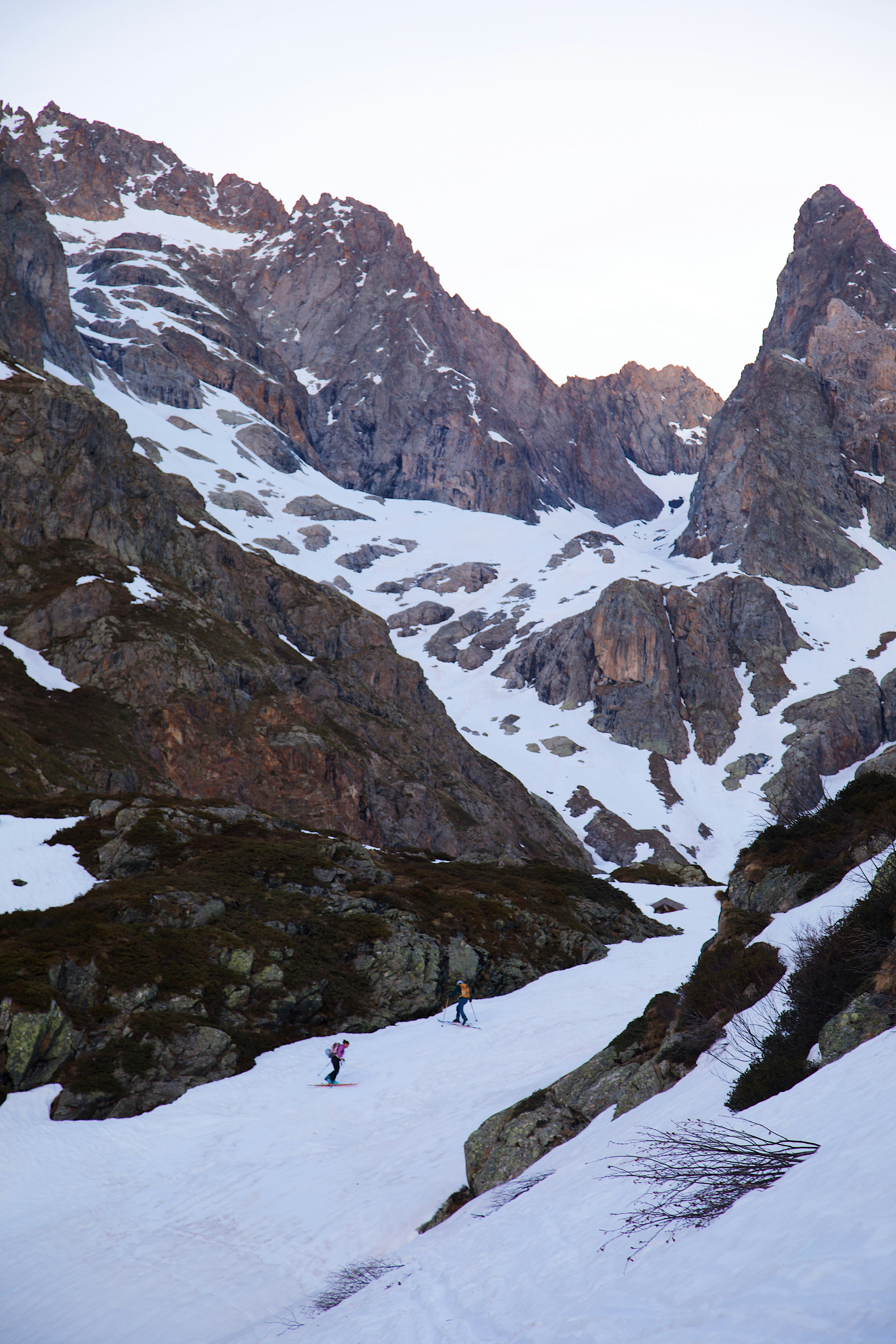Two people backcountry skiing on a snow-covered mountain slope below rocky peaks