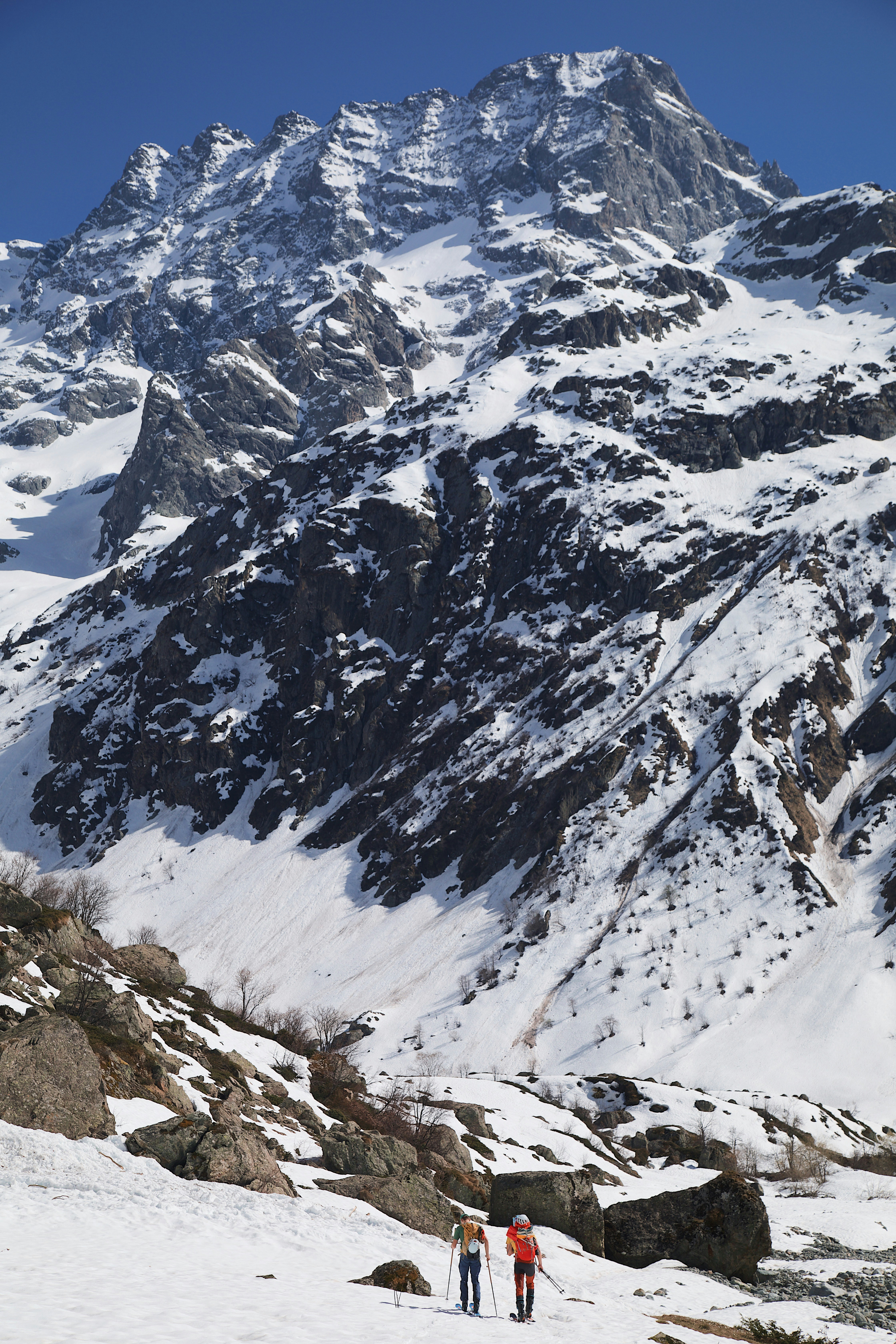 Two people hiking on a snow-covered mountain slope below a rocky peak