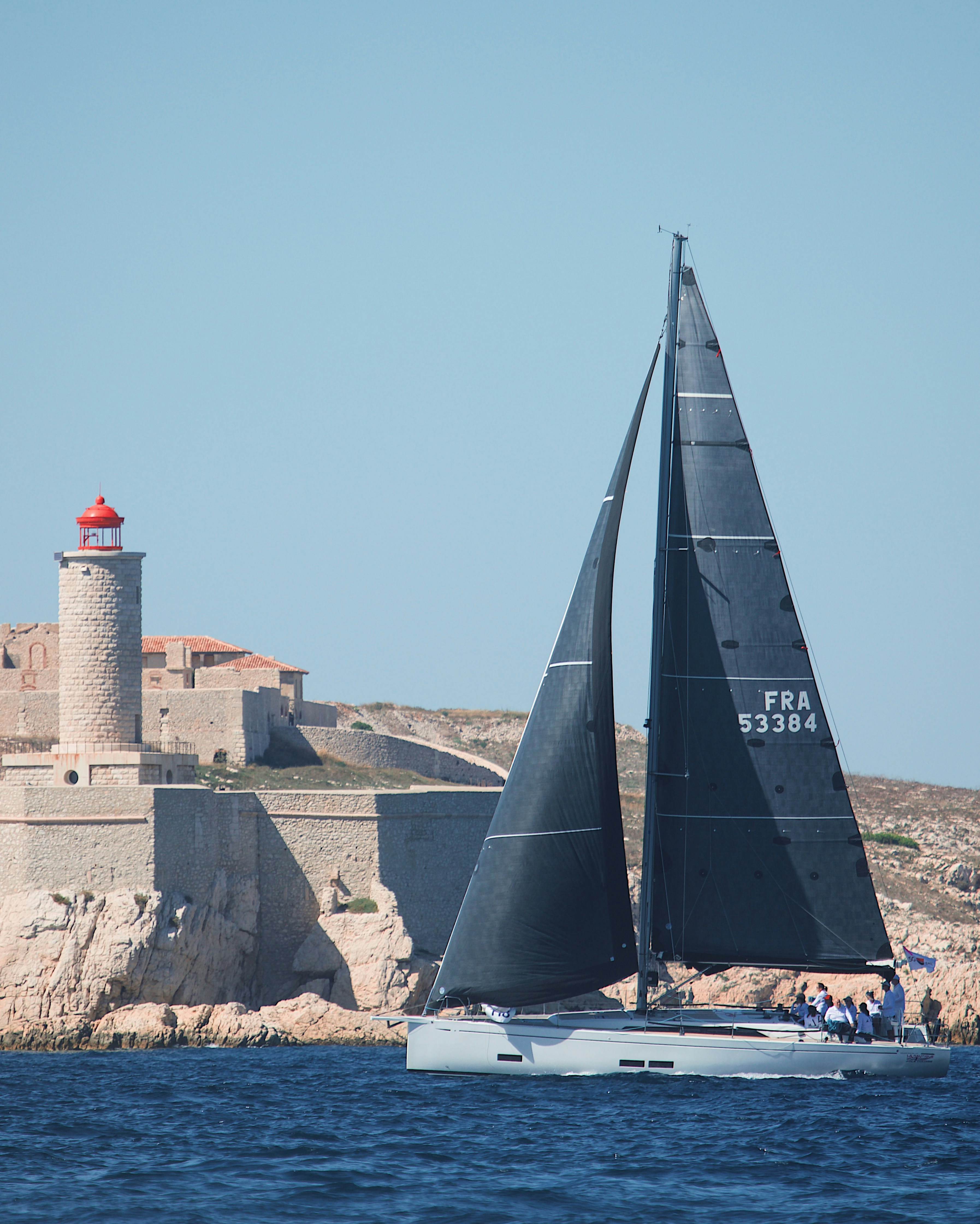 A sailboat with black sails passing a stone lighthouse on a rocky coast