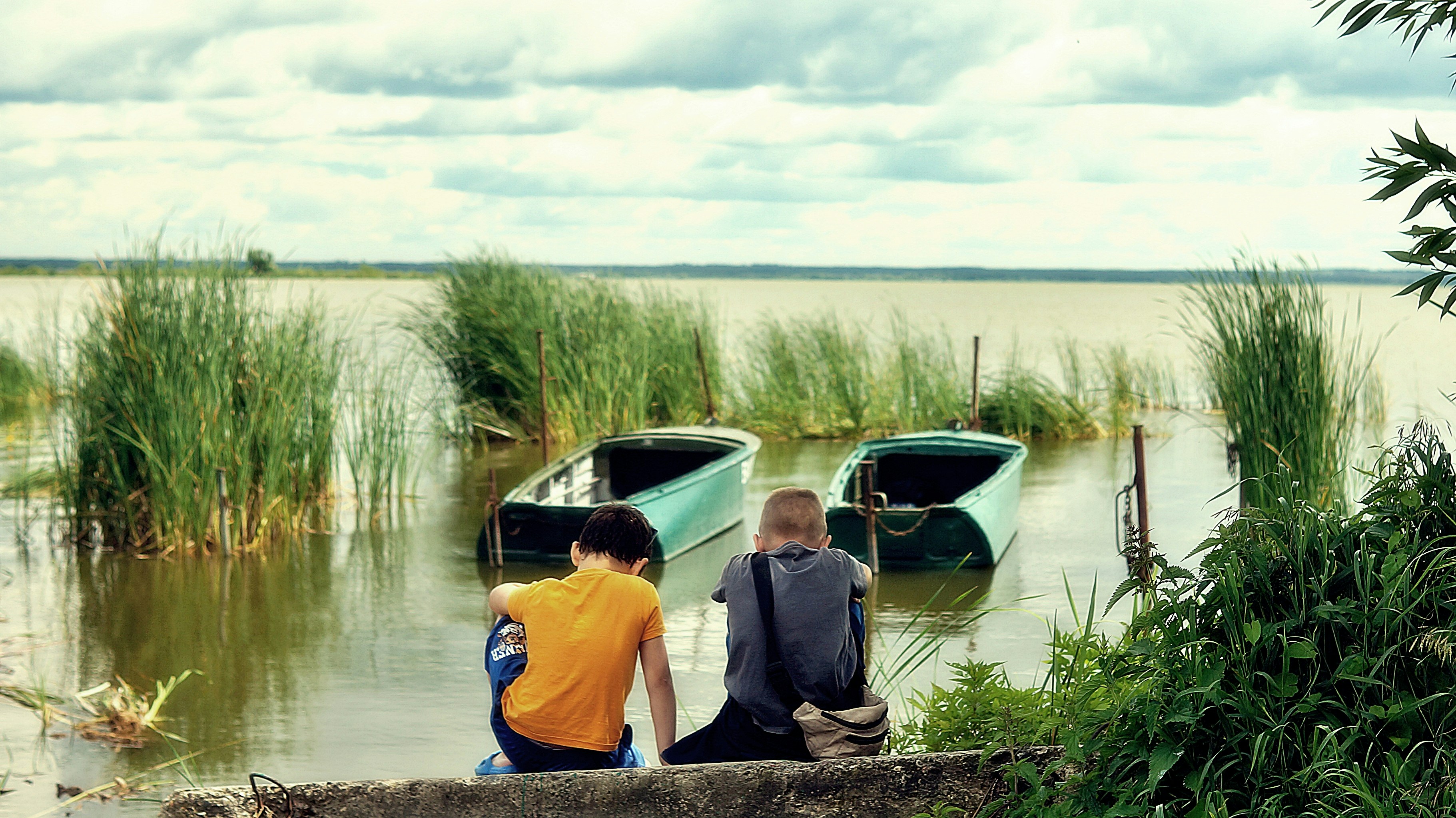 Two children sitting on a concrete ledge overlooking a lake with two green boats
