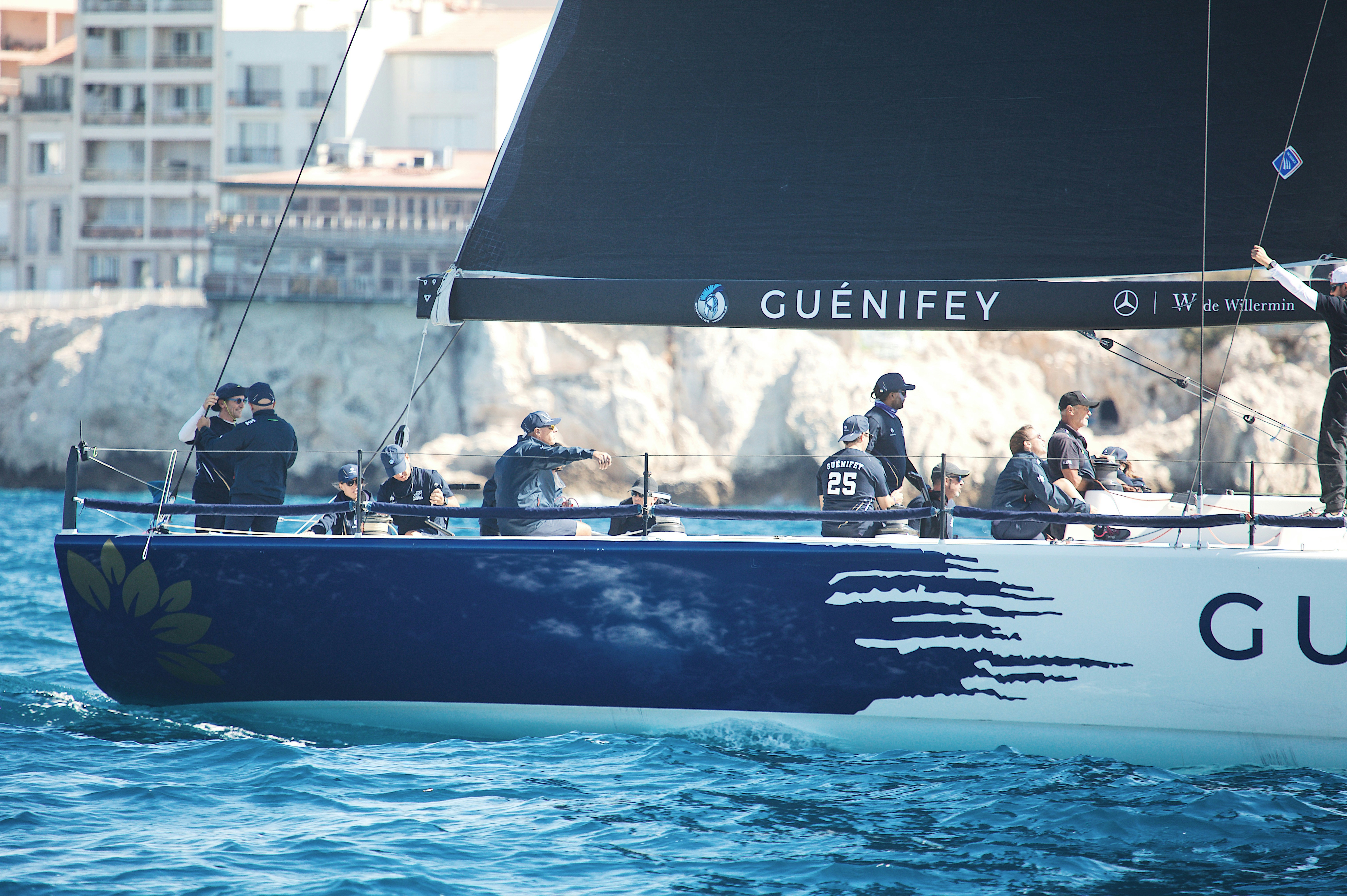 A crew in matching dark uniforms sailing a racing yacht near a rocky coastline