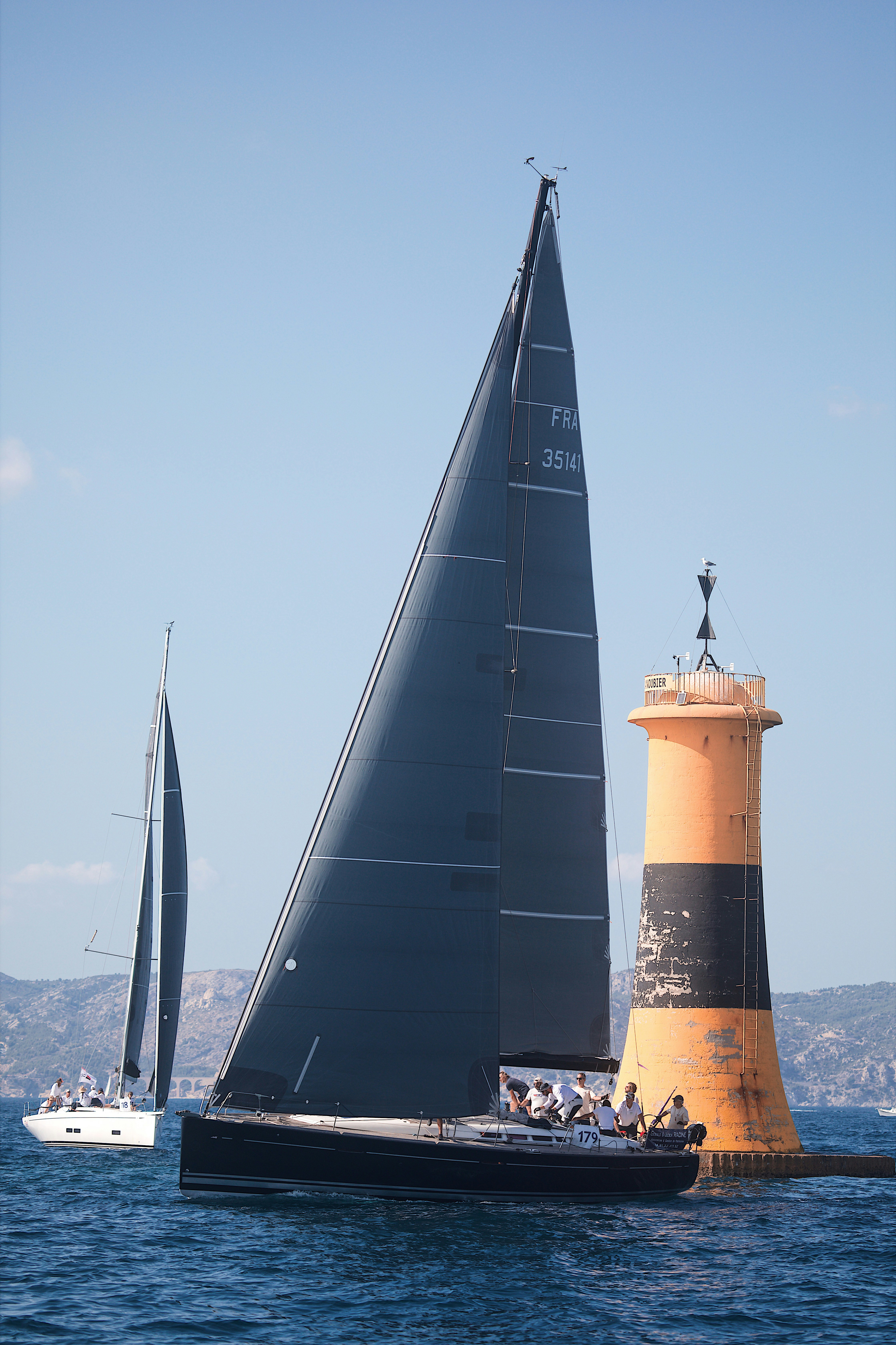 A black sailboat passing a yellow and black lighthouse on the blue sea