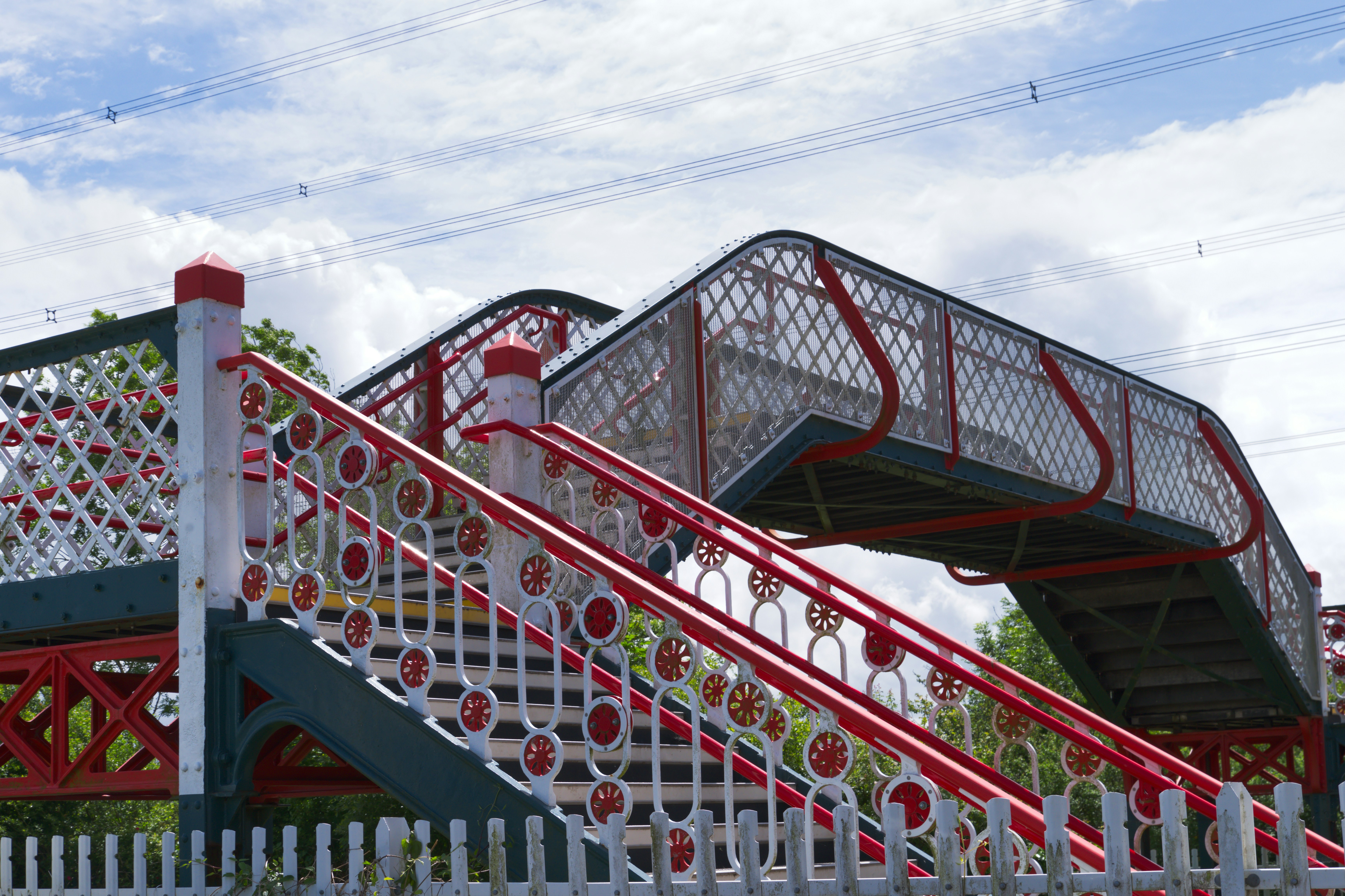 A Victorian style pedestrian footbridge with red and white metal railings against a cloudy sky
