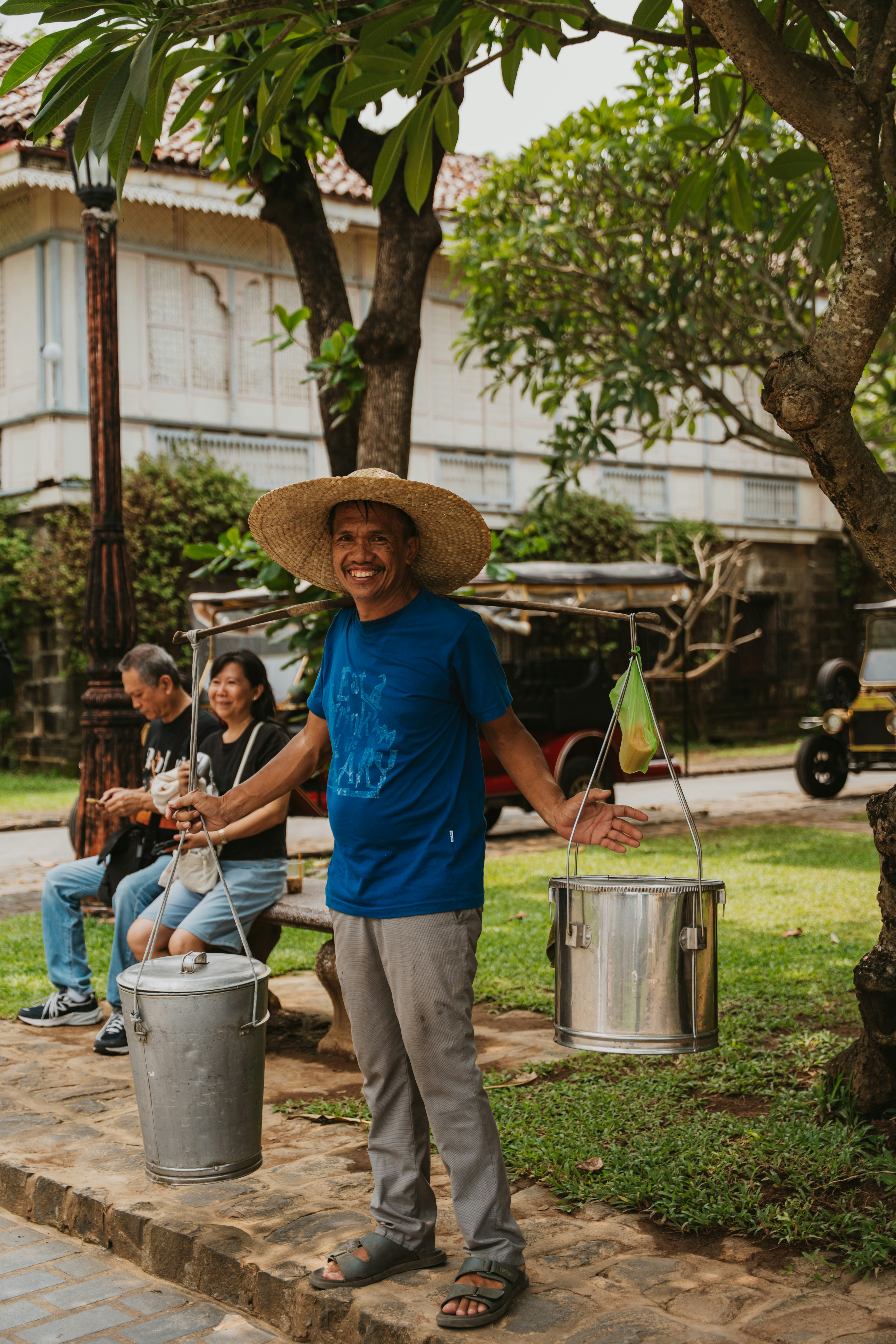 A smiling street vendor in a straw hat carrying metal containers on a yoke