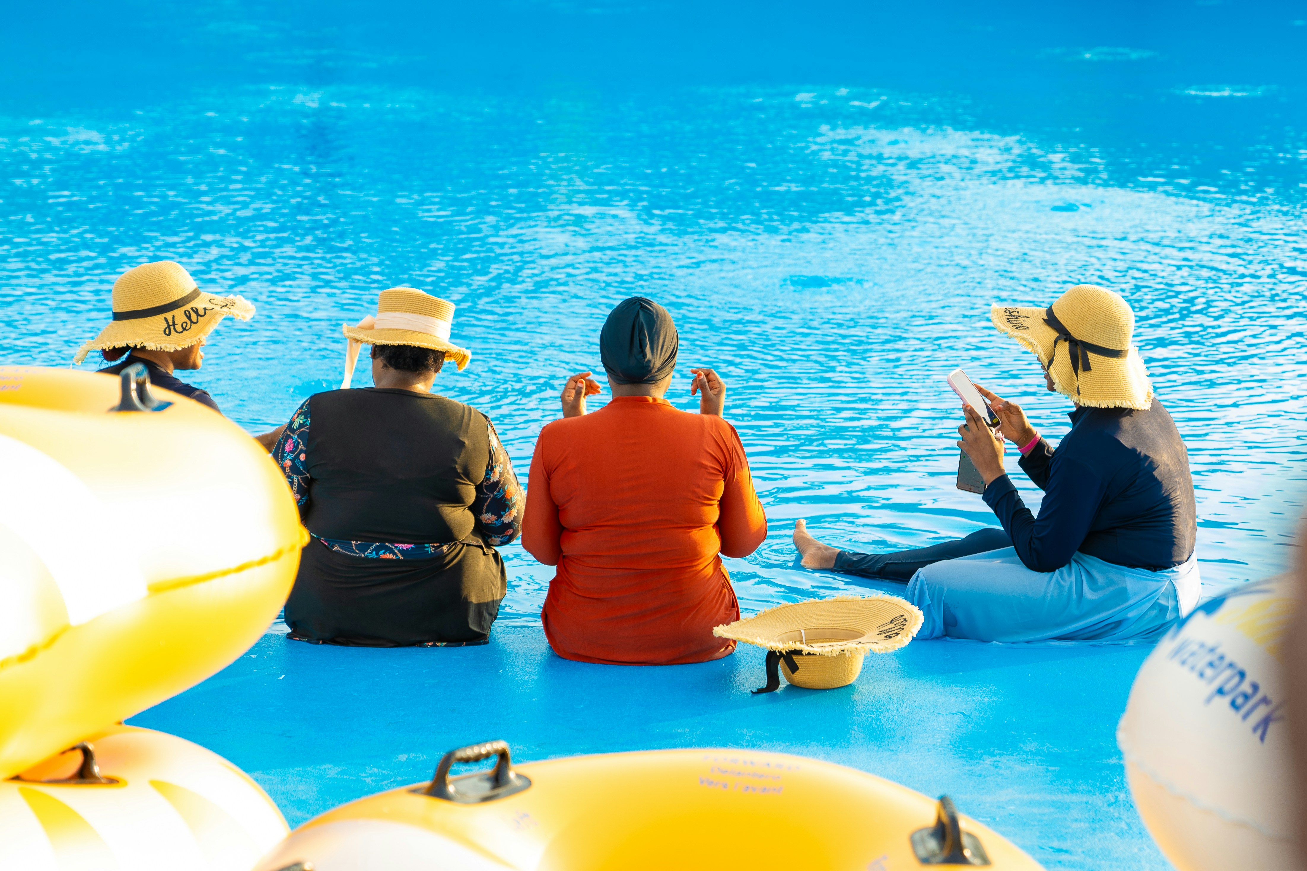 Four people in swimwear and sun hats sitting at the edge of a pool