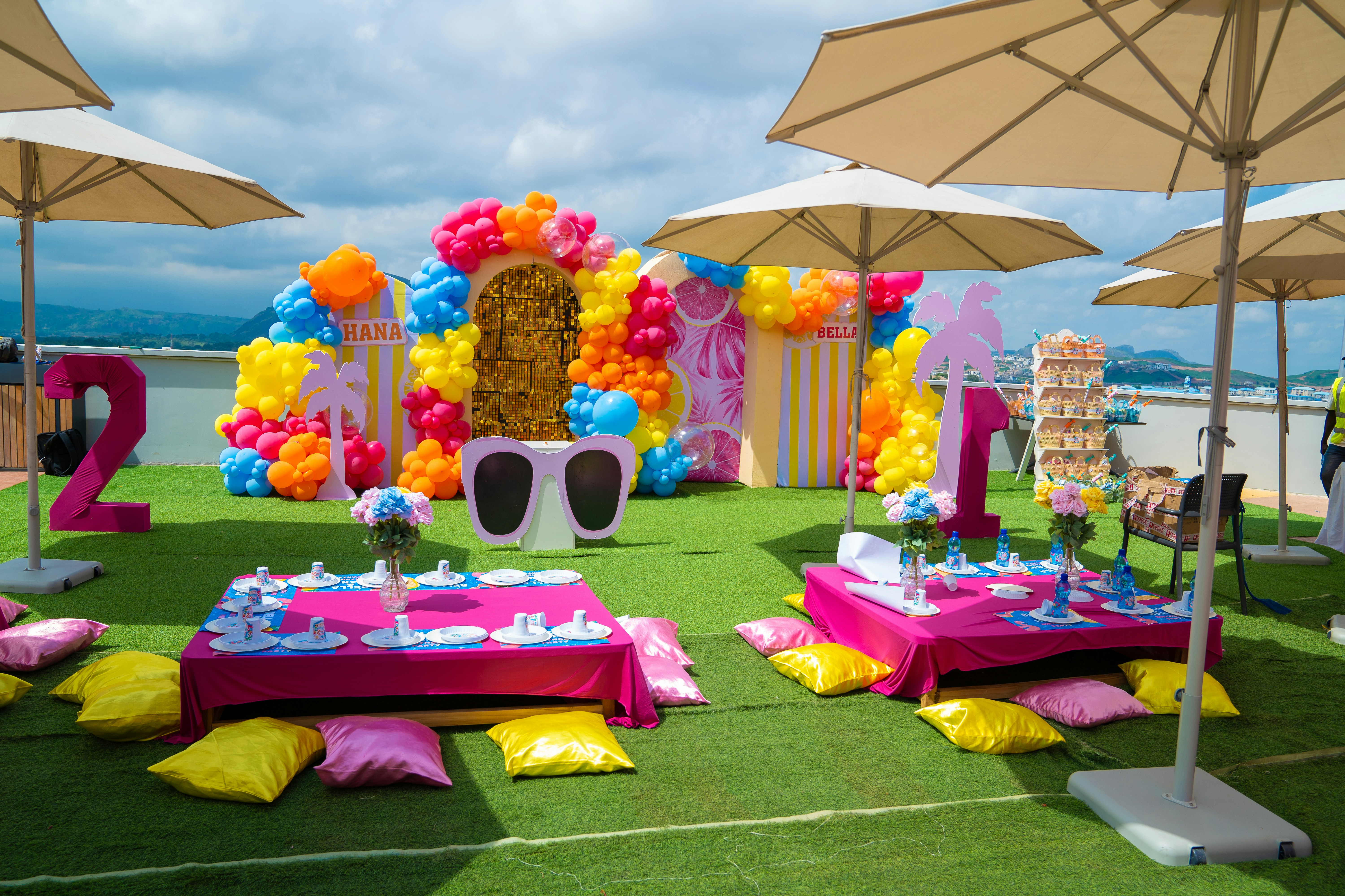 Outdoor party setup with low pink tables, colorful balloons, and umbrellas on grass