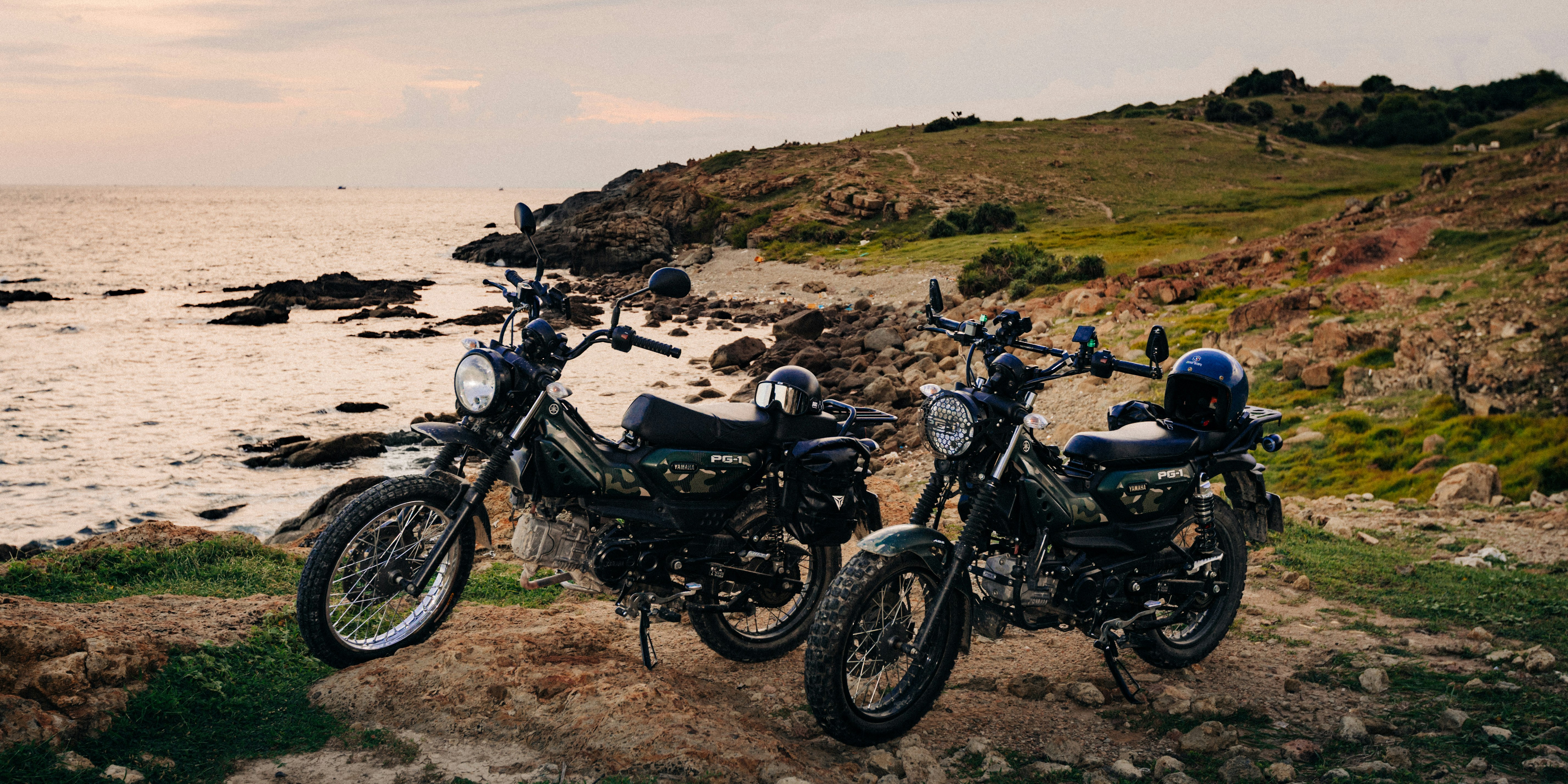 Two dark green motorcycles parked on a rocky coastal path at golden hour