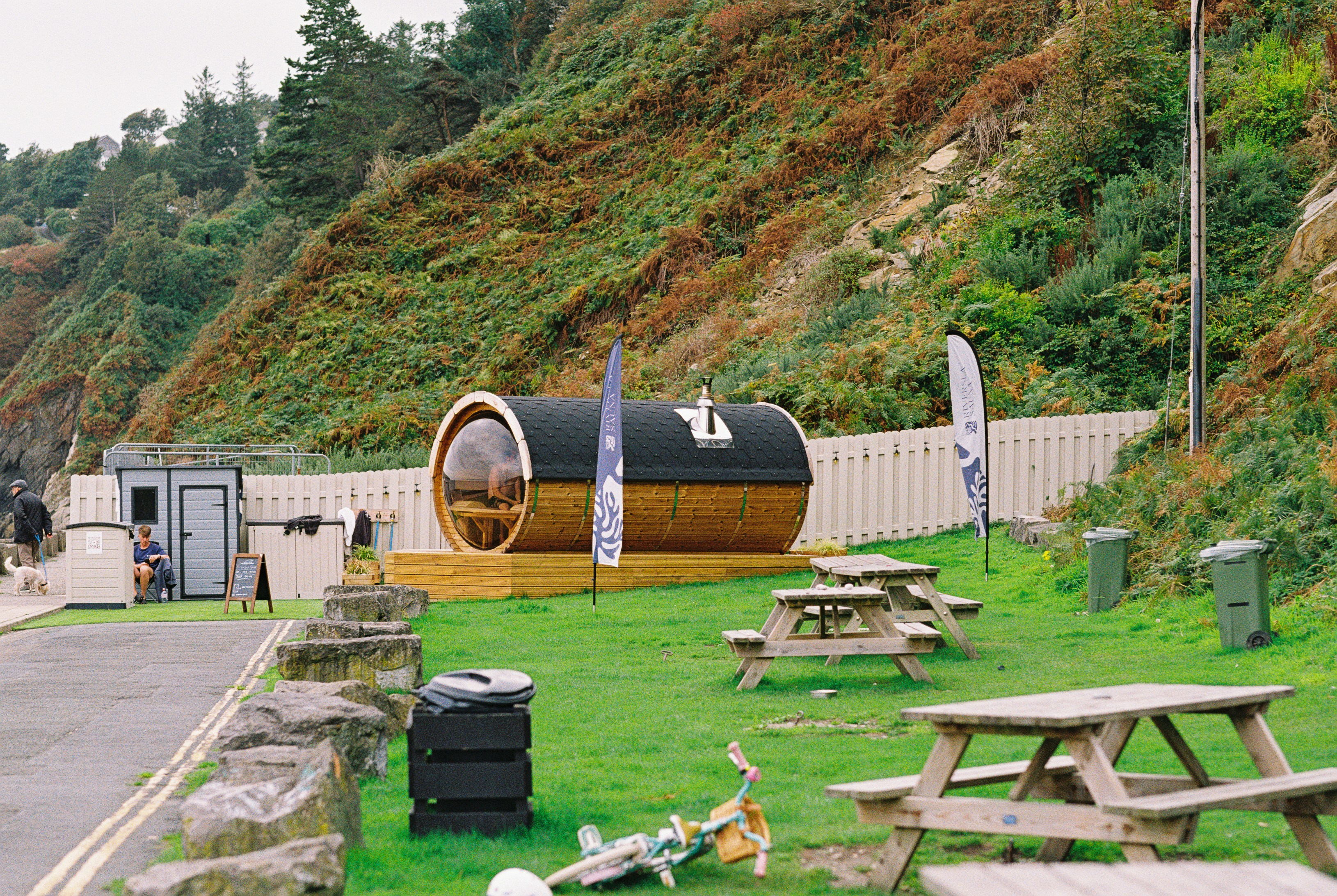 Wooden barrel sauna on a grassy lawn below a hillside