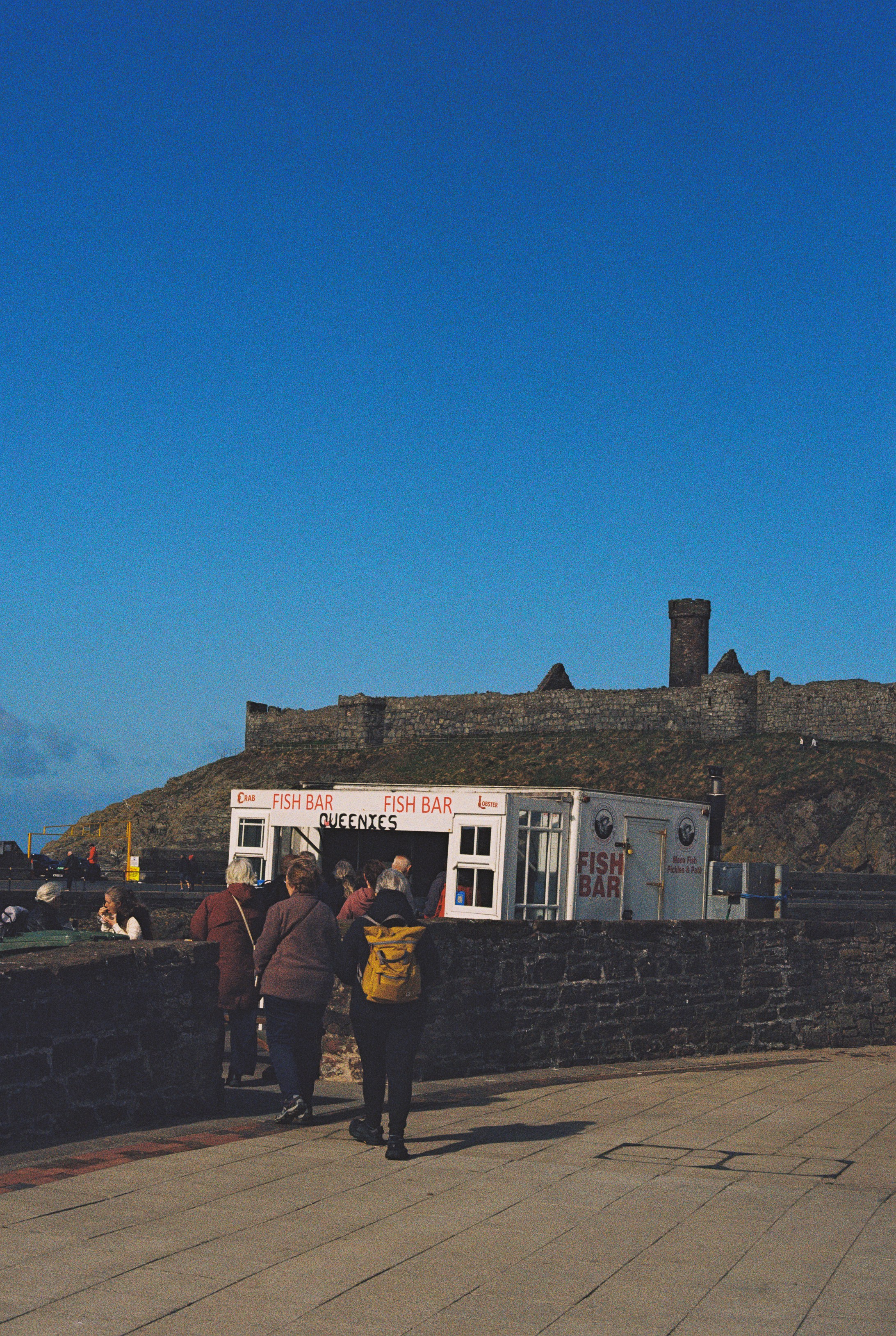 People walking toward a fish bar kiosk below a stone castle ruin