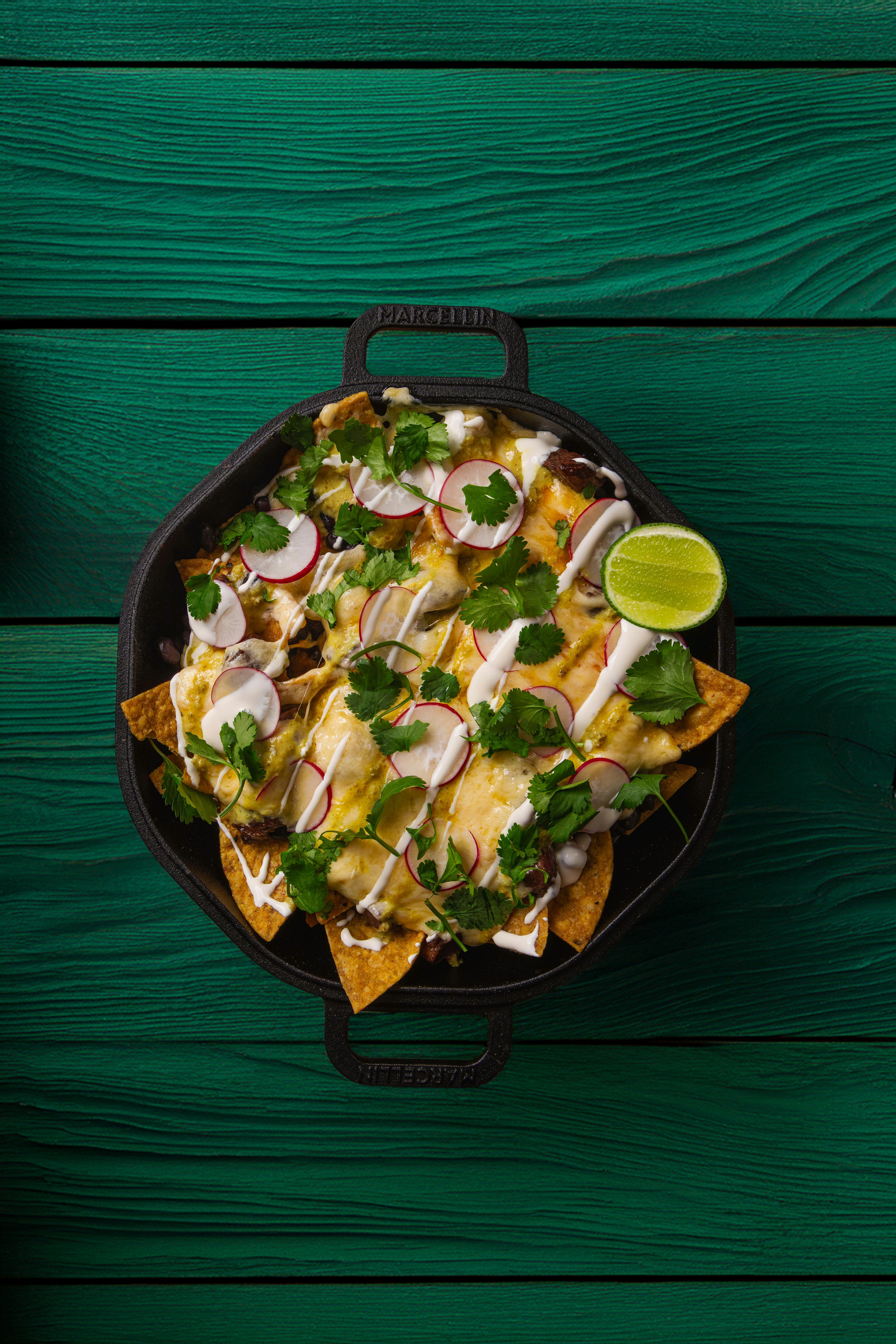 Nachos with melted cheese, radish slices, cilantro, and lime on a green wooden table