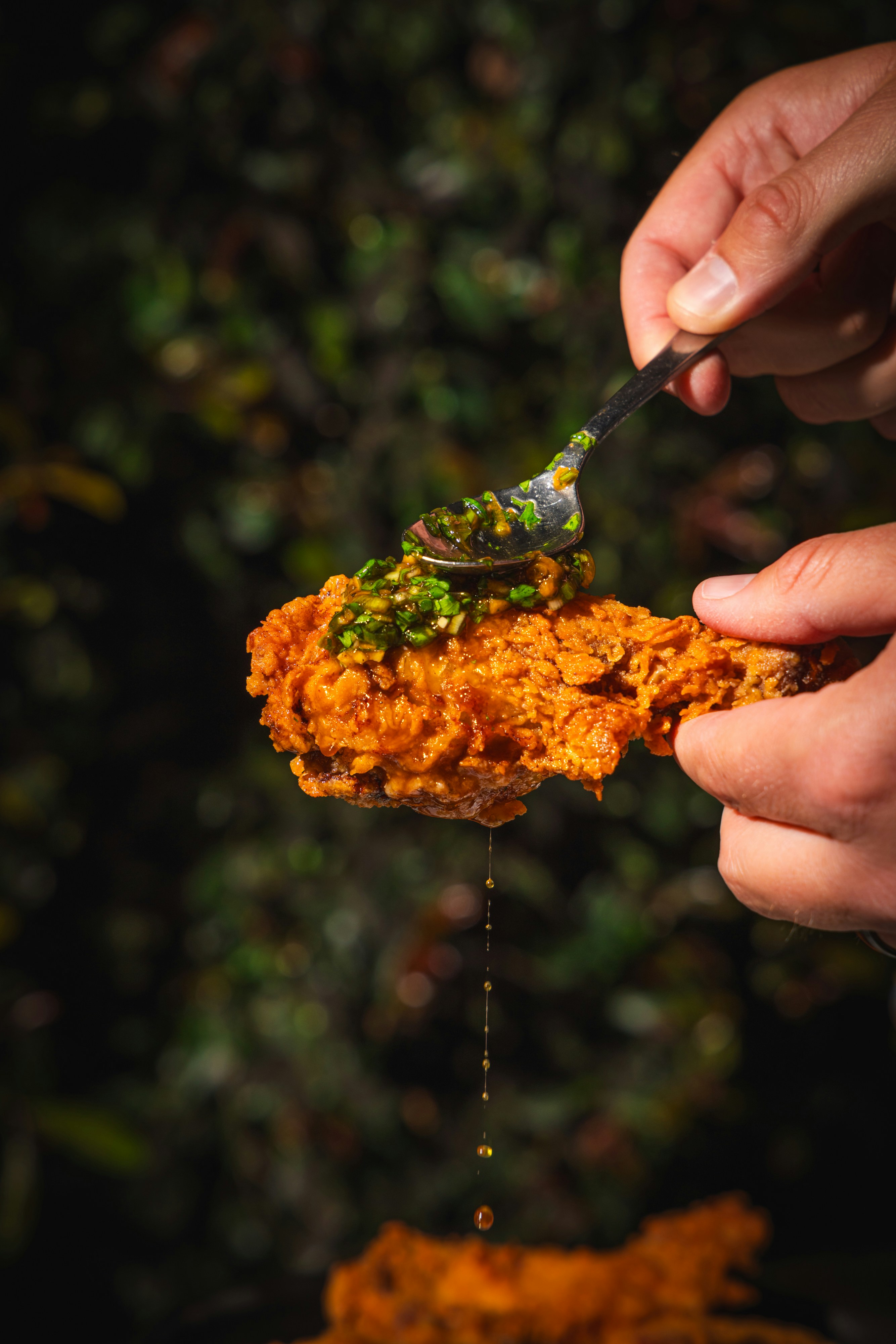 A person holding a piece of fried chicken while spooning green herb sauce over it